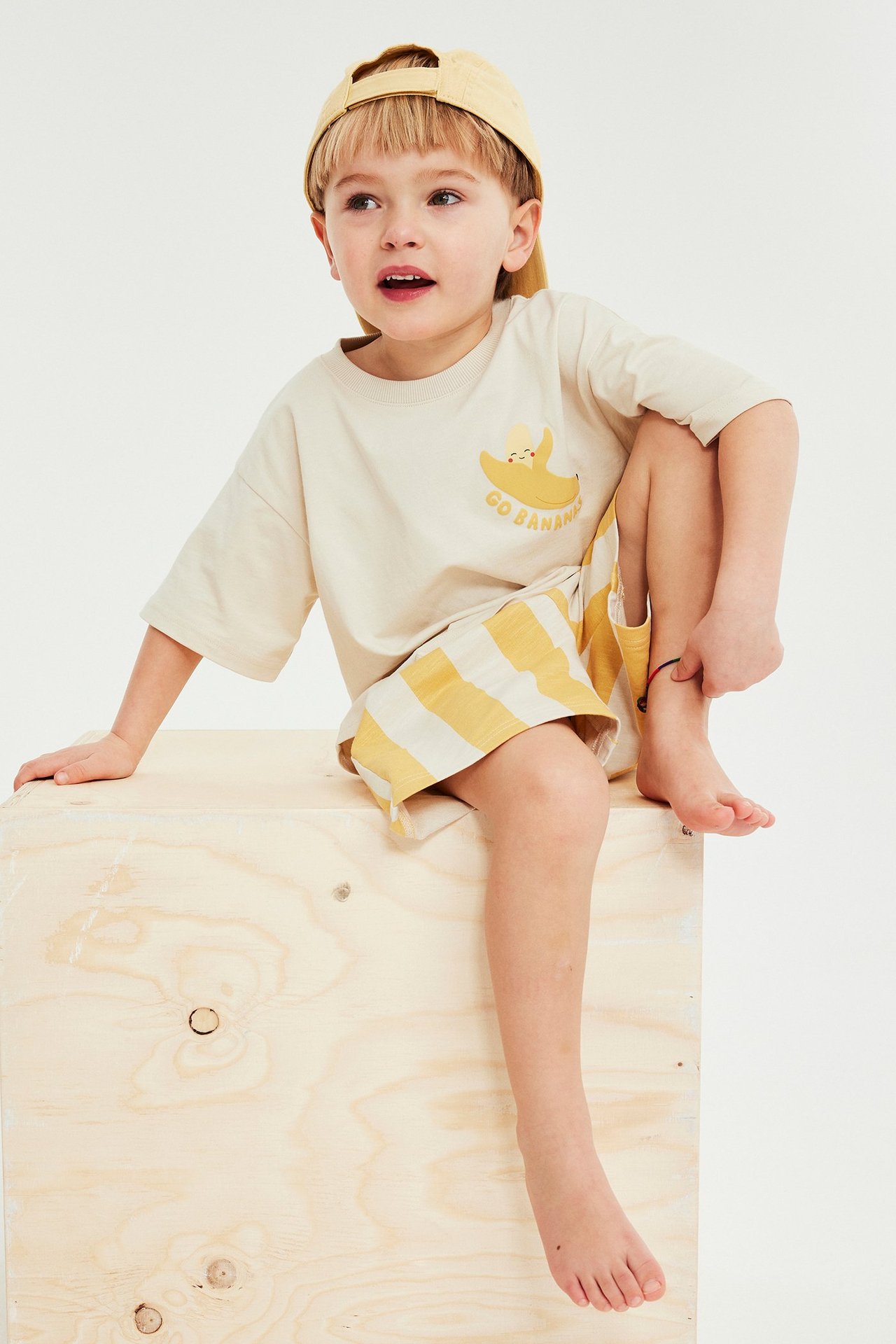 Edestä, lapsi pukeutunut beigen banaaniprintti-T-paitaan, kelta-valkoraidallisiin shortseihin ja keltaiseen lippalakkiin.