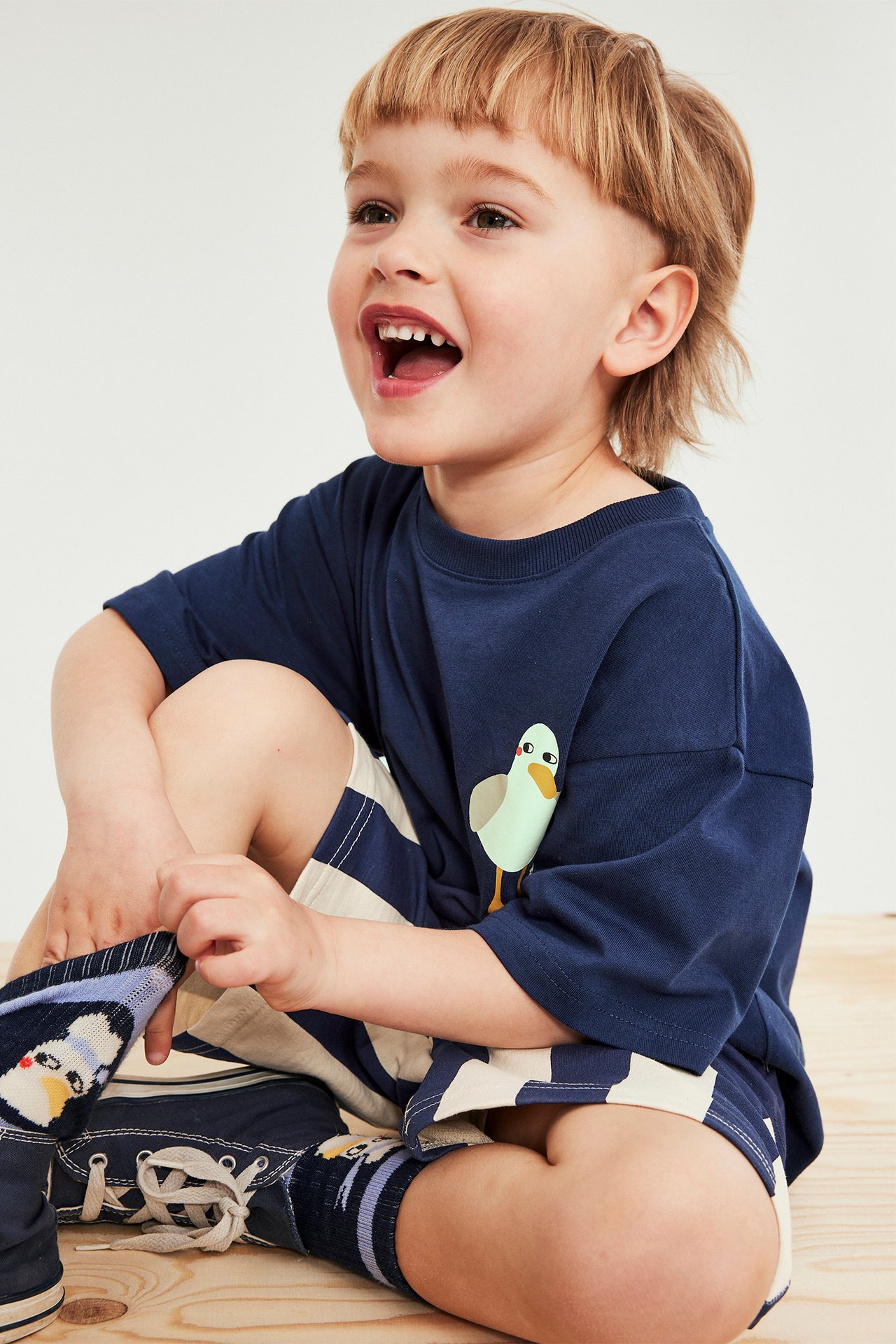 Leende barn bär blå bomulls t-shirt med måstryck, randiga shorts, mönstrade strumpor och blå skor. Frontvy.