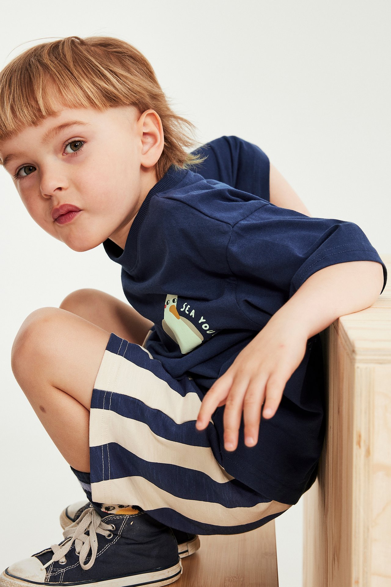 Framifrån, ett barn i mörkblå bomulls-t-shirt med tryck och randiga mörkblå och beige shorts.