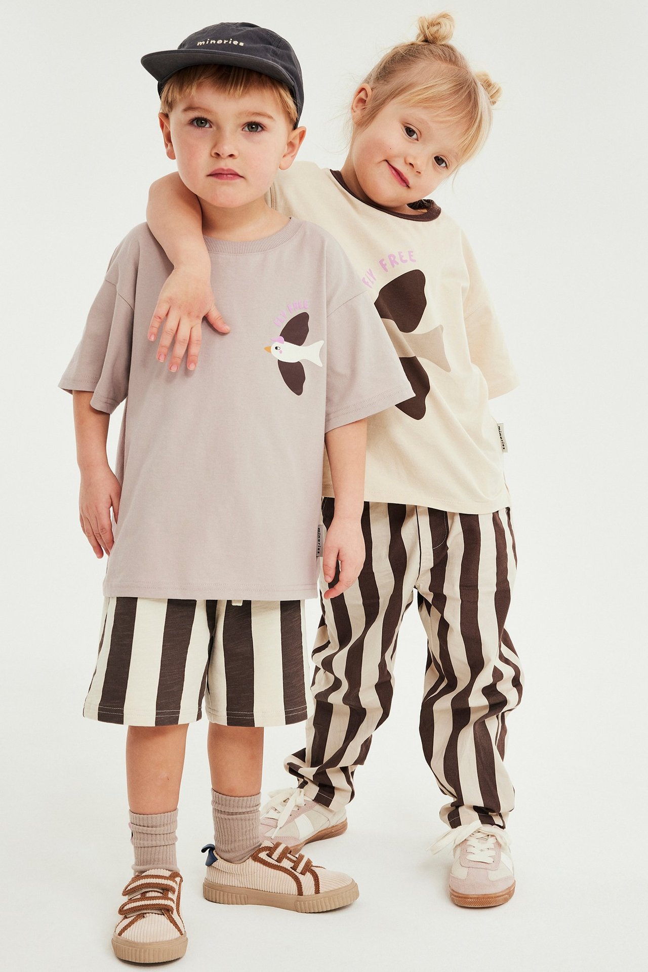 Widok z przodu: Dziecko w beżowej koszulce z nadrukiem i brązowych bawełnianych szortach w paski, drugie dziecko w spodniach w paski.