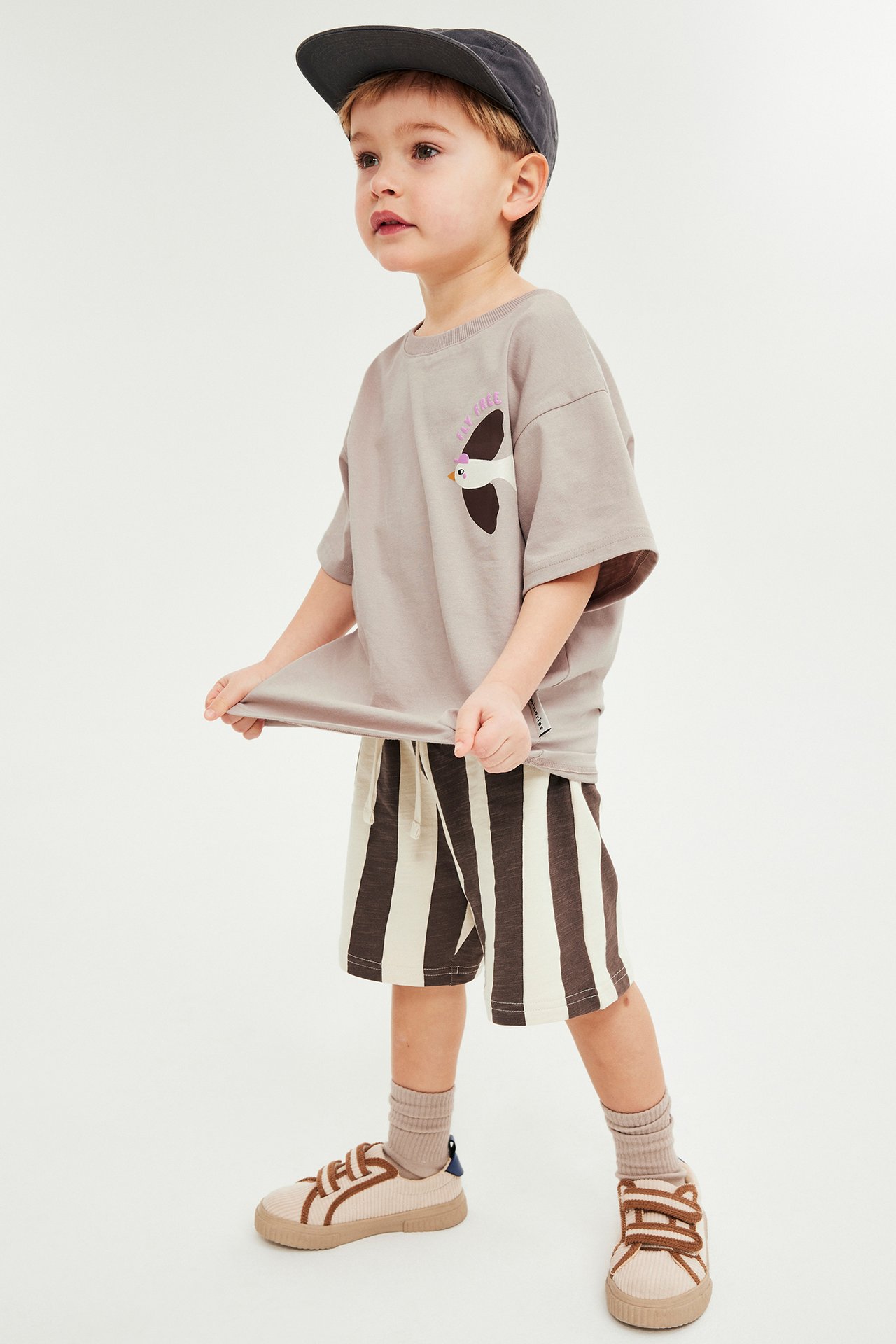 Z przodu: Dziecko w beżowym T-shircie z ptakiem, brązowych pasiatych szortach, ciemnoszarej czapce, beżowych trampkach.