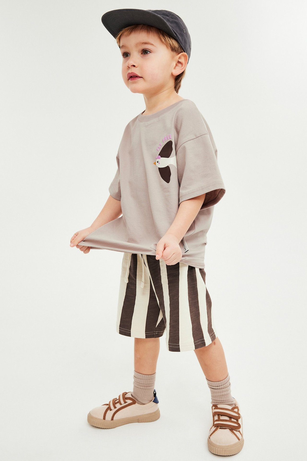 Z przodu: Dziecko w beżowym T-shircie z ptakiem, brązowych pasiatych szortach, ciemnoszarej czapce, beżowych trampkach.
