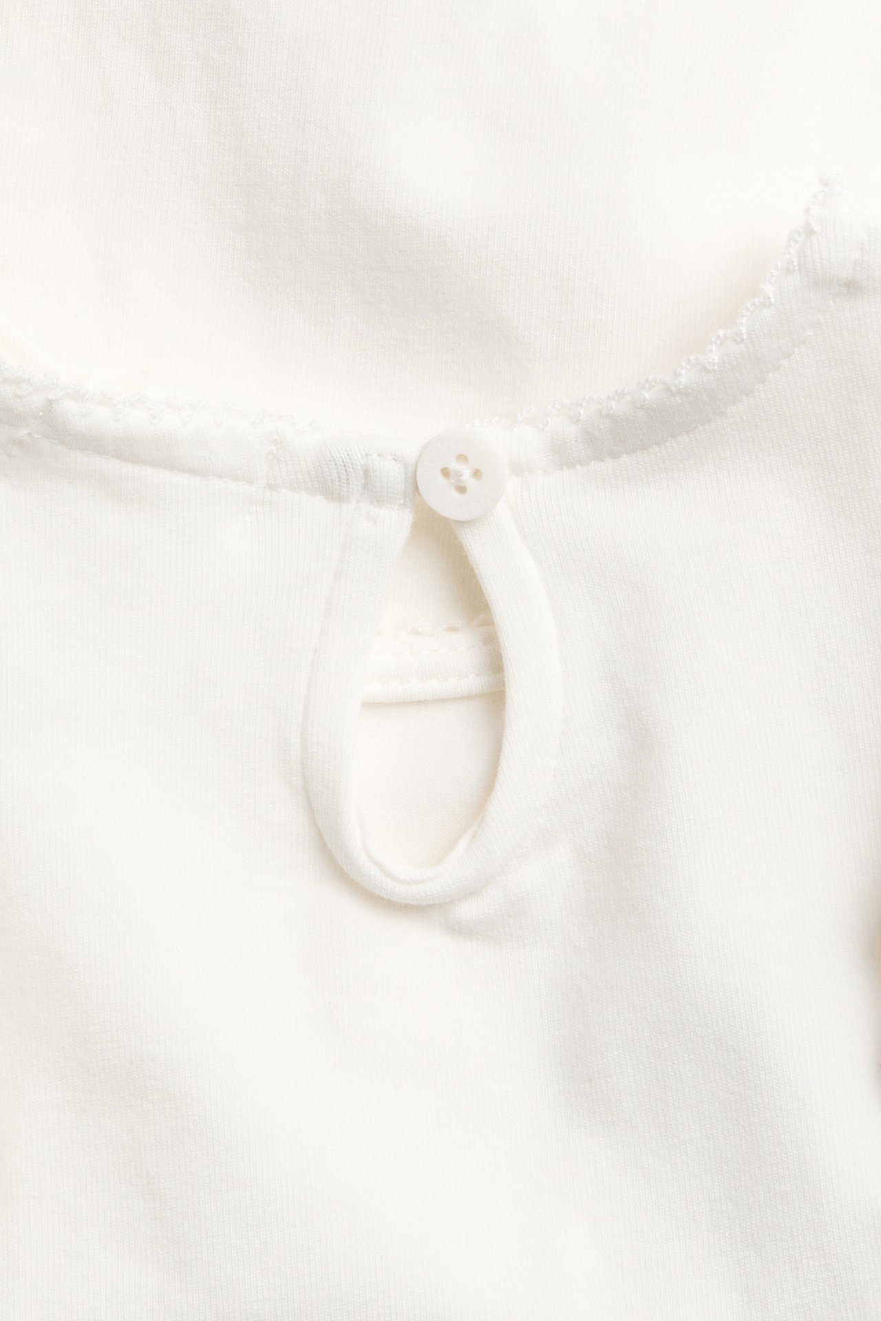 Nærbilde: Offwhite babybody i bomull, ryggutringning med nøkkelhullsåpning, knapp og picotsøm.