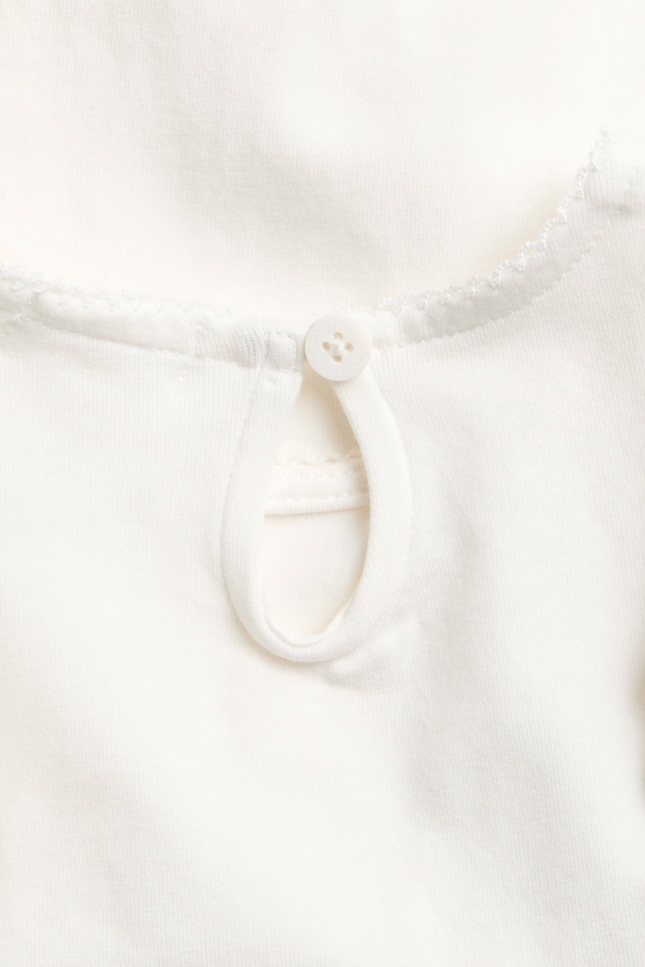 Nærbilde: Offwhite babybody i bomull, ryggutringning med nøkkelhullsåpning, knapp og picotsøm.