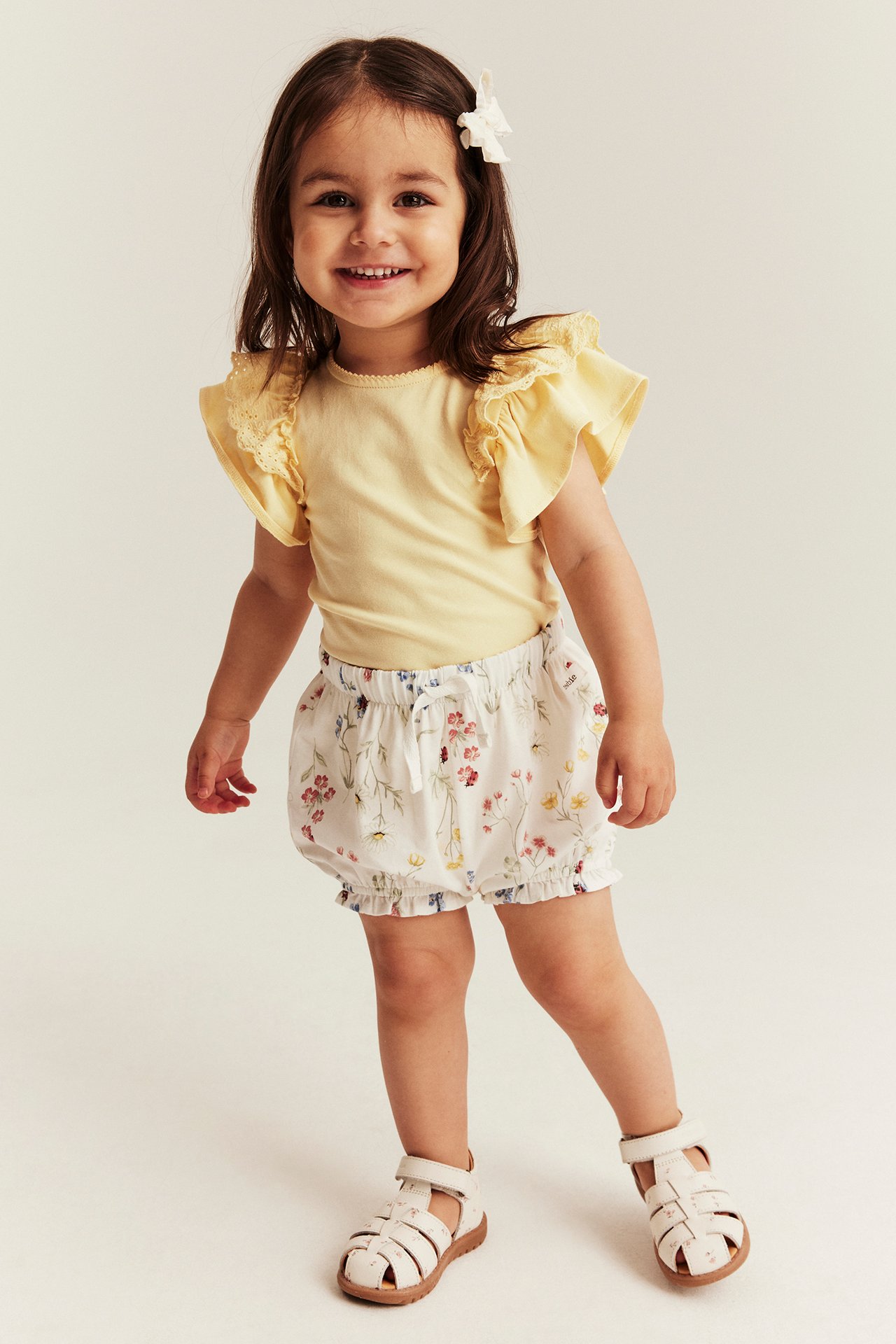 Lapsi edestä: keltainen lyhythihainen body pitsisillä röyhelöhihoilla, valkoiset kukkakuosiset shortsit ja valkoiset sandaalit.