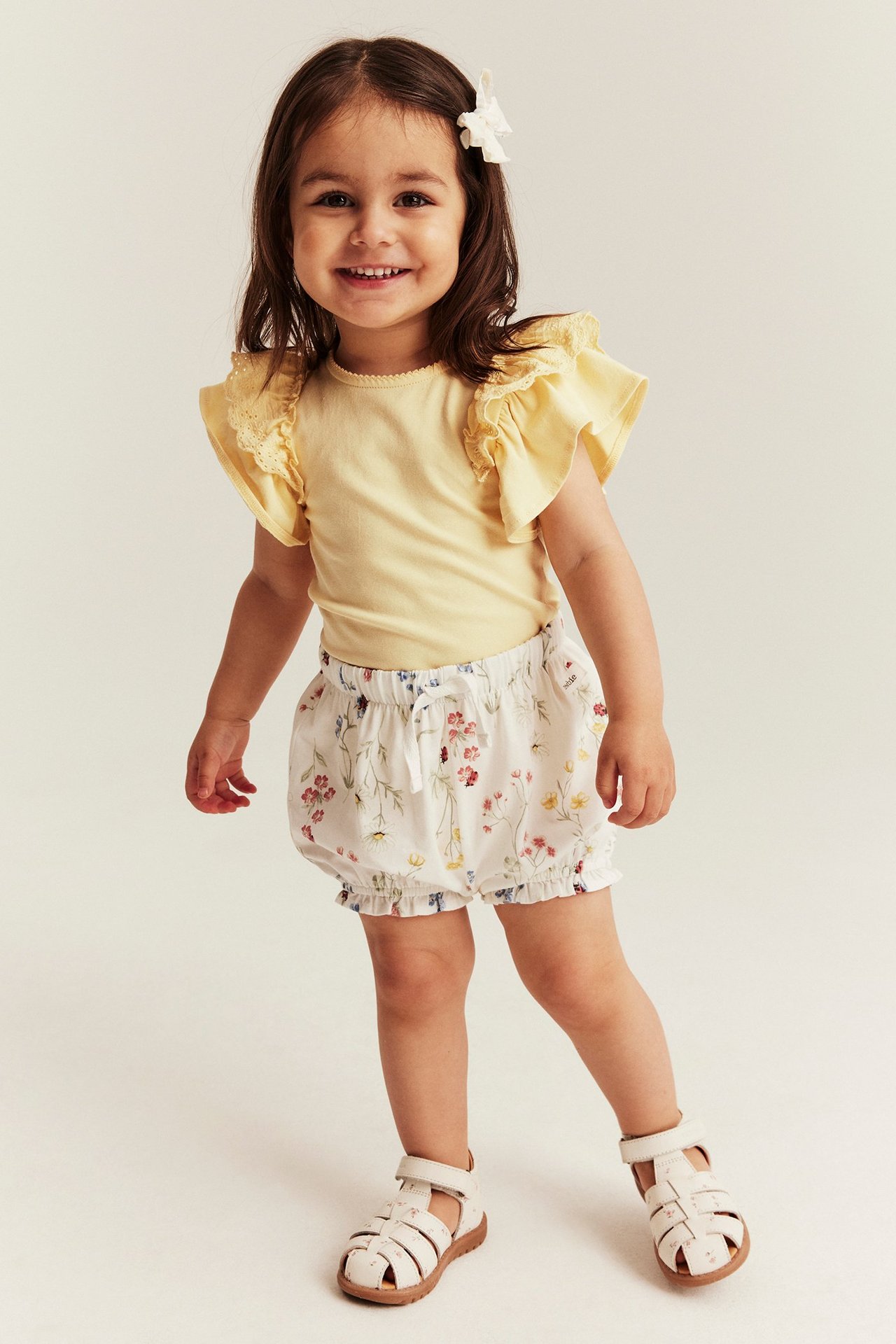 Frontbilde av et barn i gul kortermet body med volangdetaljer, hvite blomstrete bloomer-shorts og hvite sandaler.