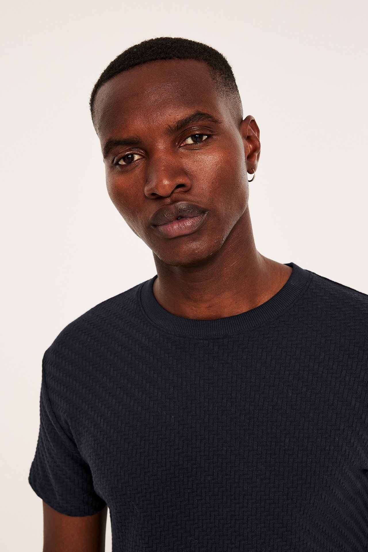 Närbild framifrån på en man i en svart texturerad bomulls-t-shirt och en liten ringörhänge.