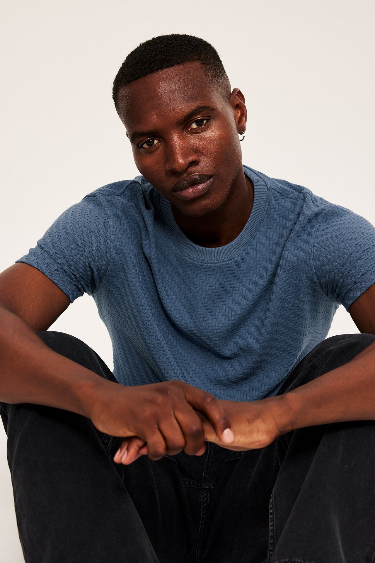 Frontvy av en man i en blå strukturerad bomulls-t-shirt, mörka byxor och en ringörhänge.