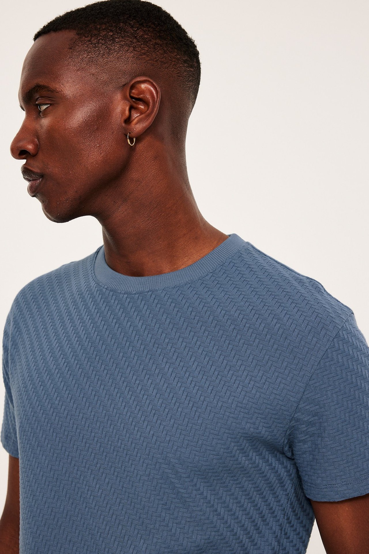 Widok z boku mężczyzny w niebieskiej bawełnianej koszulce ze wzorem strukturalnym i złotym kolczykiem.