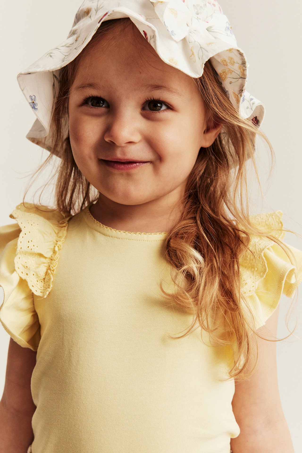 Närbild, framifrån, av ett barn i gul kortärmad topp med spetsvolanger och en blommig solhatt.