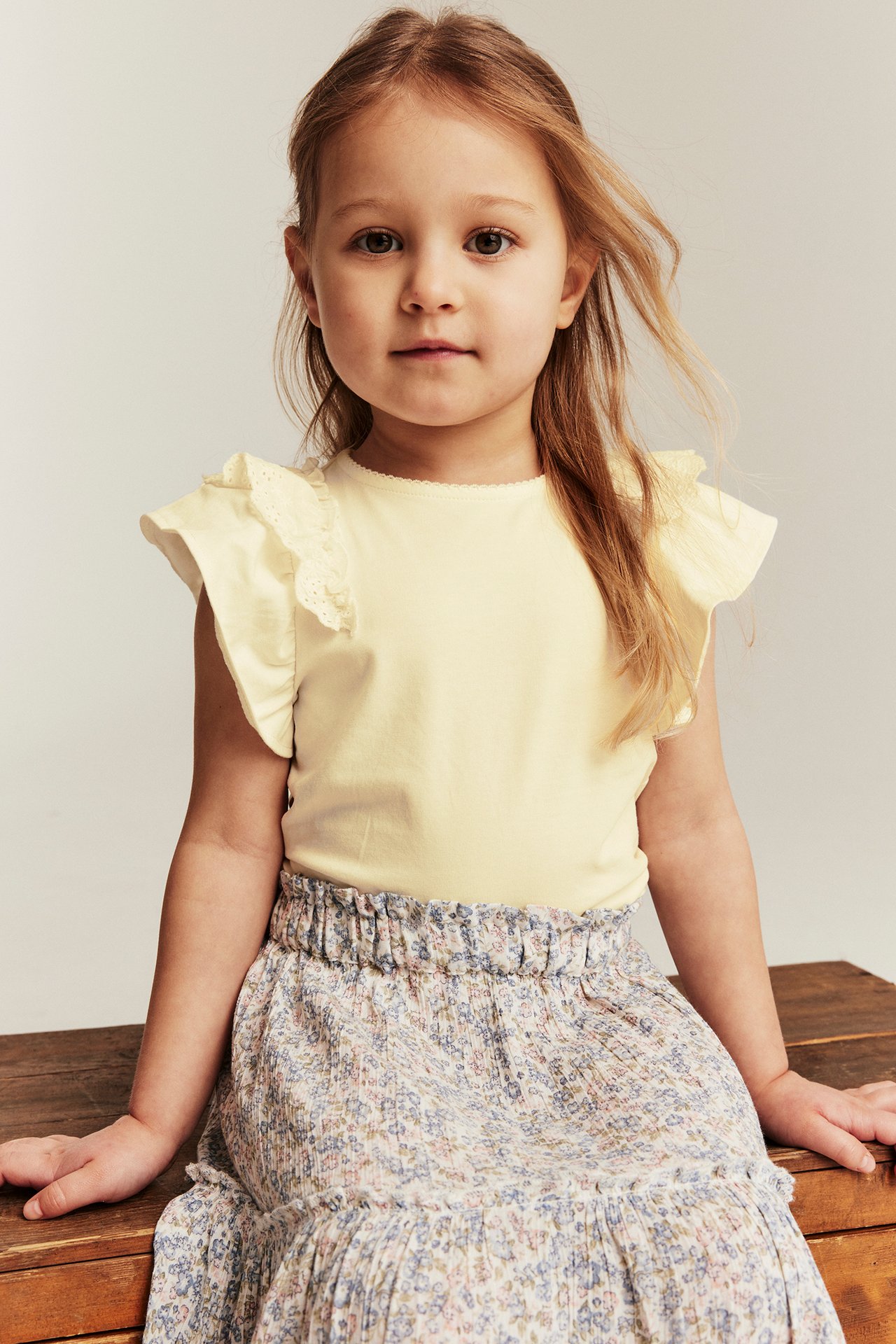Forfra: Et barn i gul kortermet topp med blondevolanger og en blomstret skjørt.
