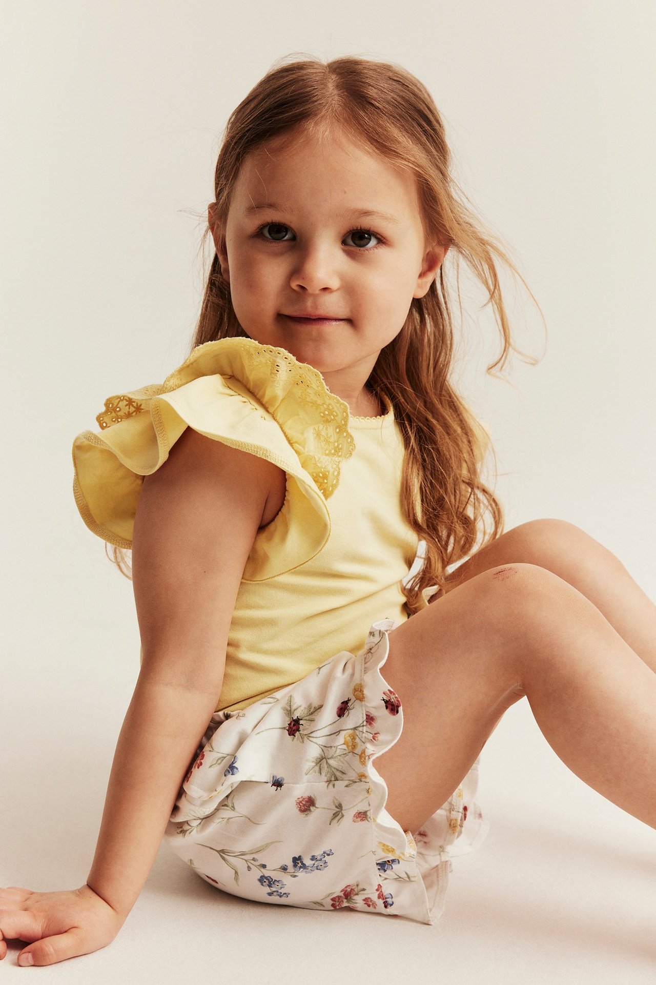 Framifrån: en unge i gul kortärmad topp med volangärm och vita, blommiga volangshorts.