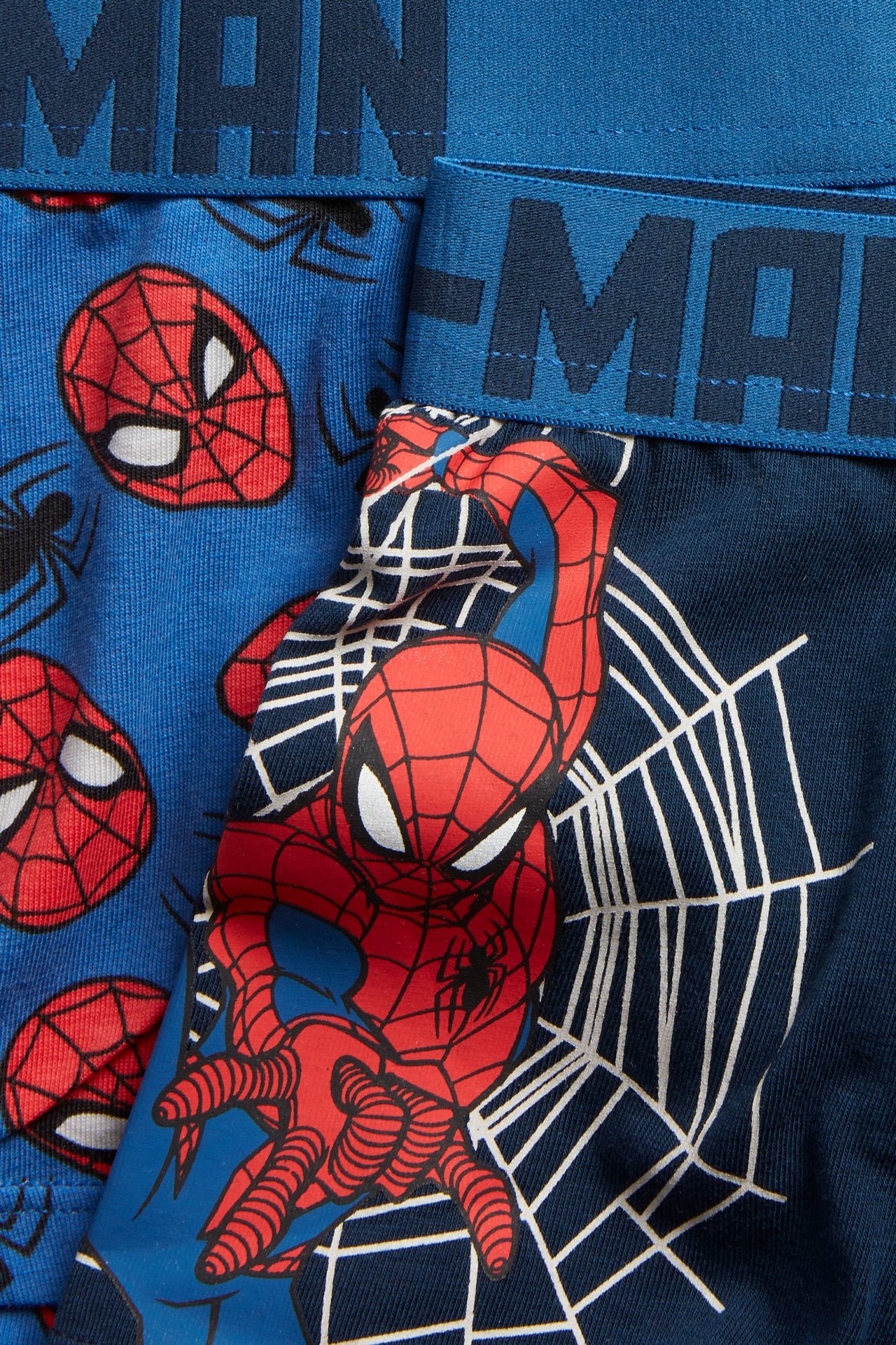 Zbliżenie z przodu na dwie niebieskie bokserki Spidermana dla dzieci, jedne we wzór maski, drugie z siecią.