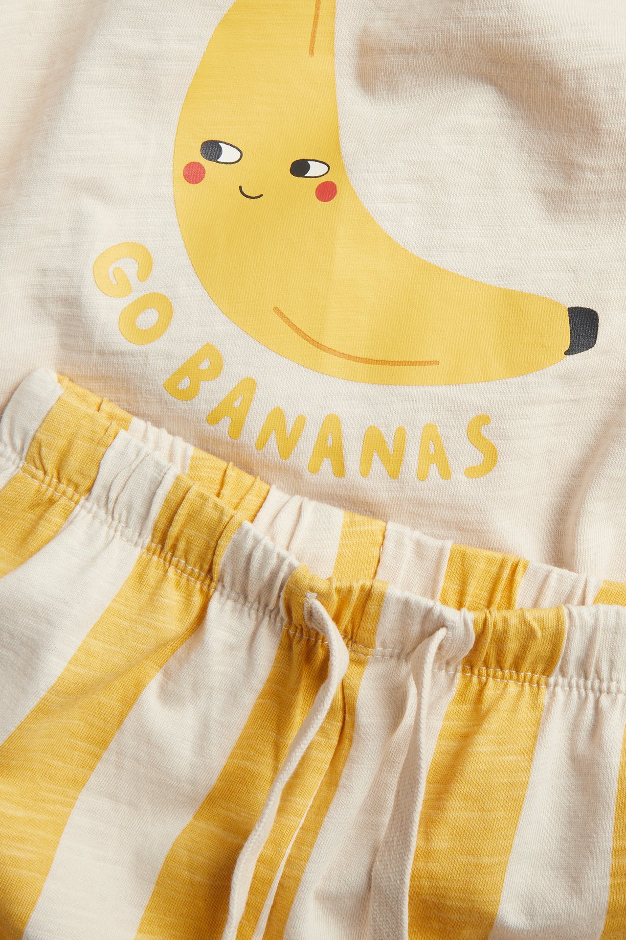 Nærbilde av et gult baby bomullssett: banantrykt t-skjorte og stripete joggebukse.