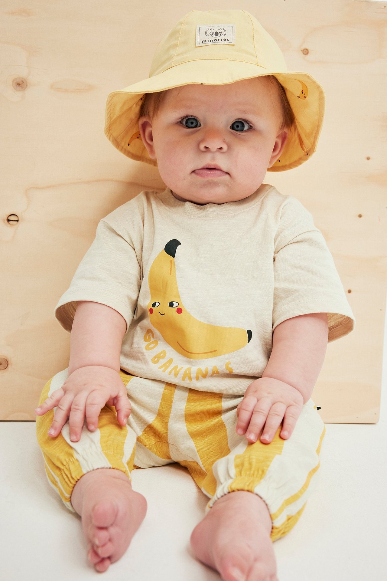 Forfra: Baby i en kremfarget T-skjorte med banantrykk, stripete gule joggebukser og en lys gul bøttehatt.