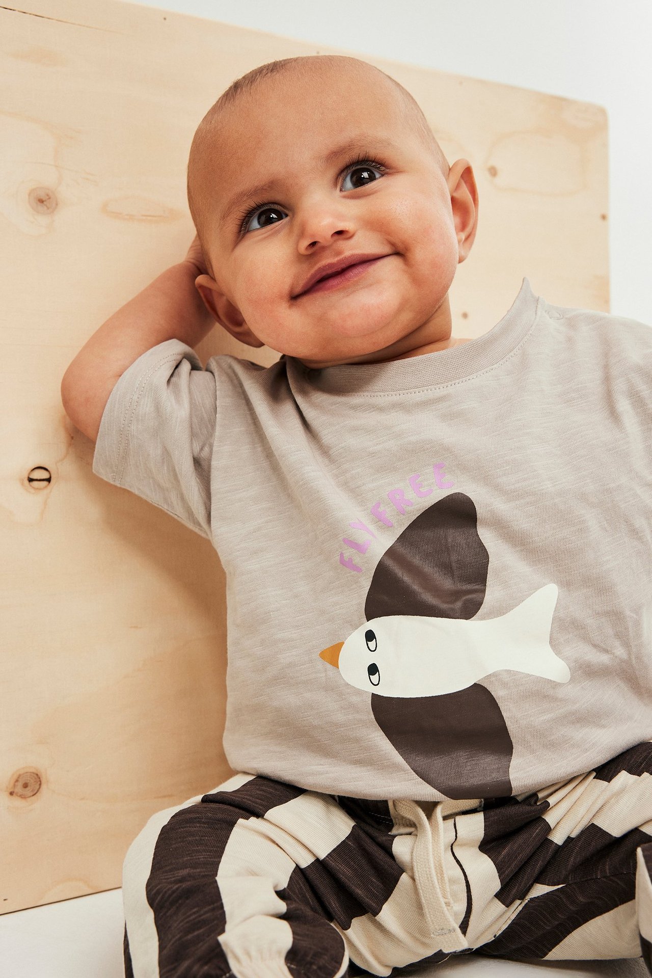 Forfra: baby i lys beige T-skjorte med fugletrykk og brunt/beige stripete joggebukse.