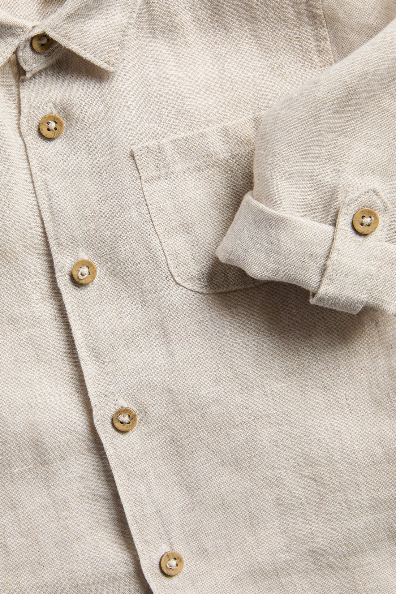Zbliżenie na jasnobeżową lnianą koszulę z kołnierzykiem, guzikami, kieszonką na piersi i podwiniętym rękawem.