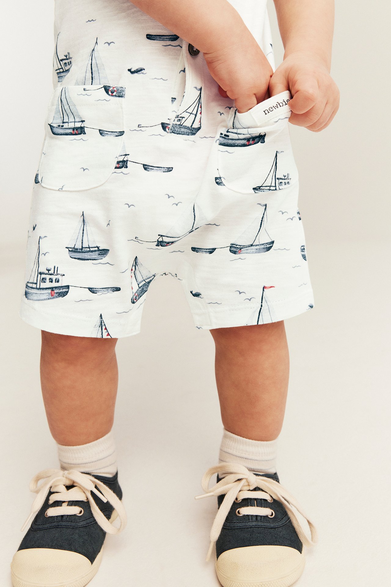 Frontvy: baby i offwhite slubtrikå-romper med segelbåtsmönster, påsydda fickor och mörkblå skor.