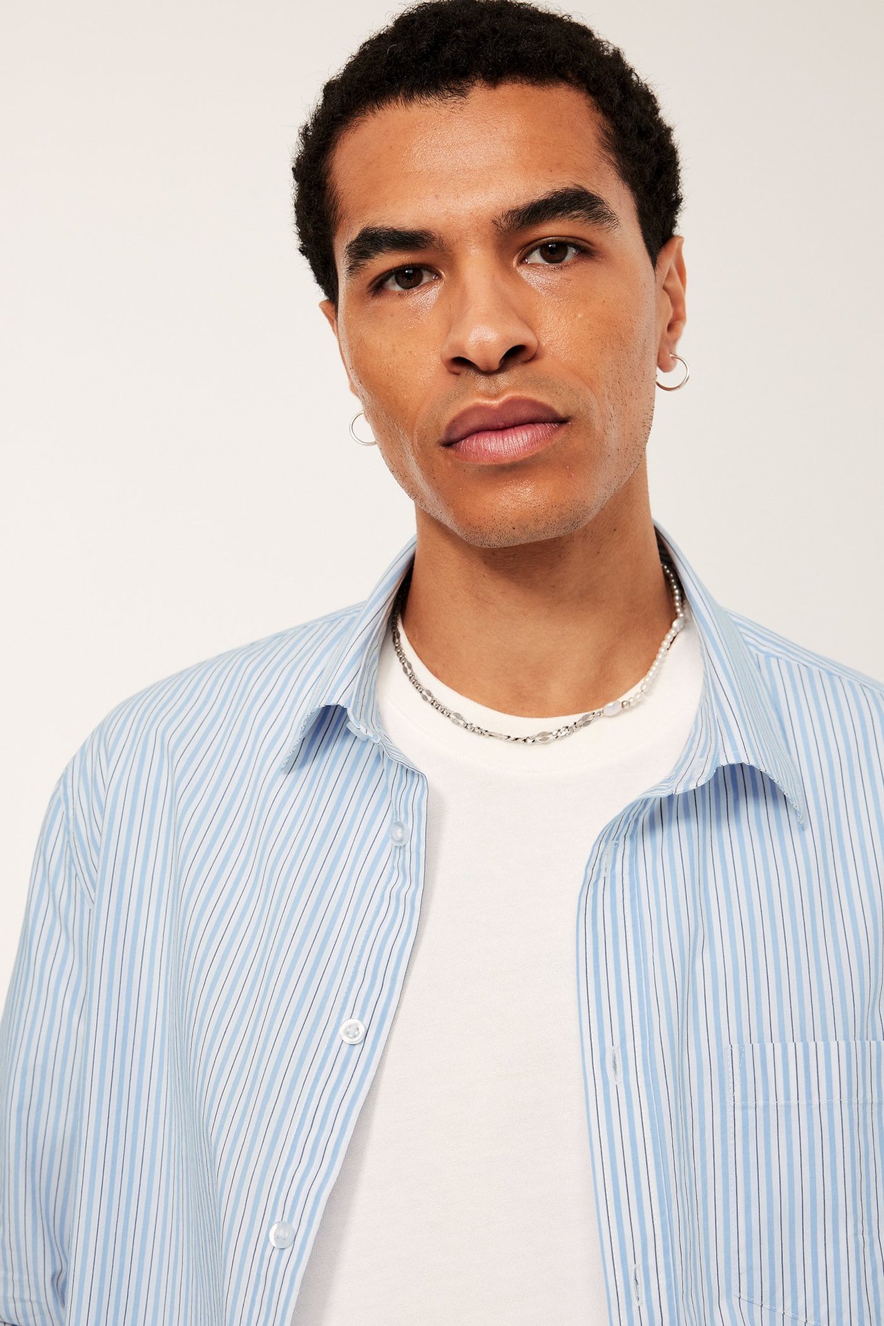 Framifrån: Man i öppen ljusblå randig poplinskjorta över vit t-shirt, med halsband och örhängen.