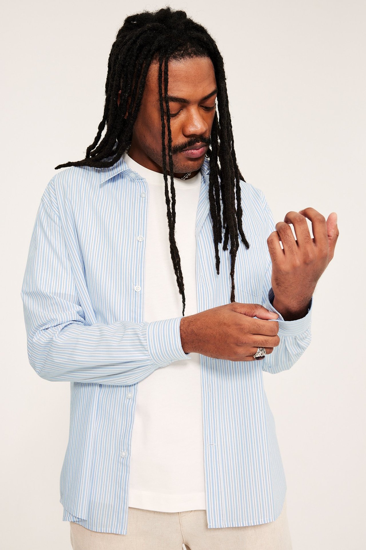 Framifrån, man justerar manschetten på sin ljusblårandiga poplinskjorta över en vit t-shirt.