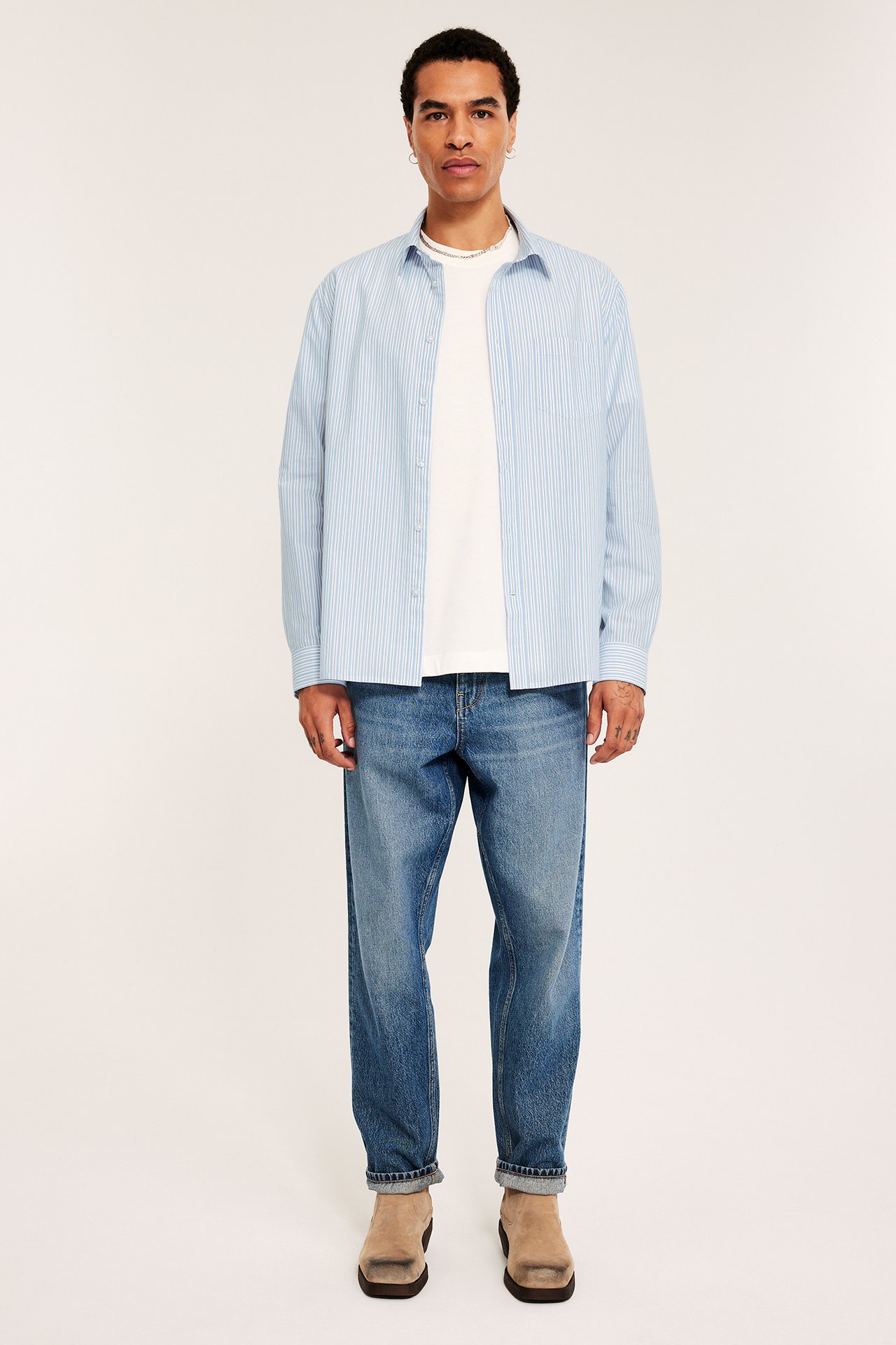 Frontvy av en man i öppen ljusblå randig poplinskjorta över vit t-shirt, blå jeans och beige boots.