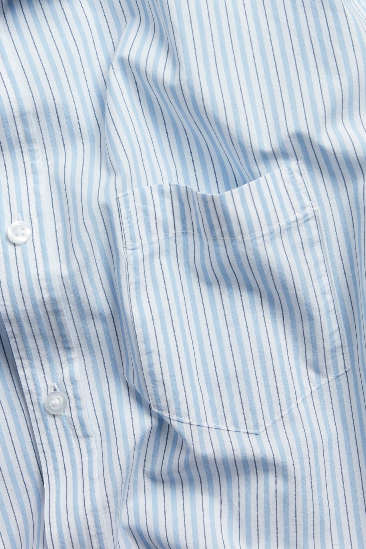 Zbliżenie koszuli popelinowej w niebiesko-białe paski, wykonanej z bawełny, z kieszenią na piersi i białymi guzikami.