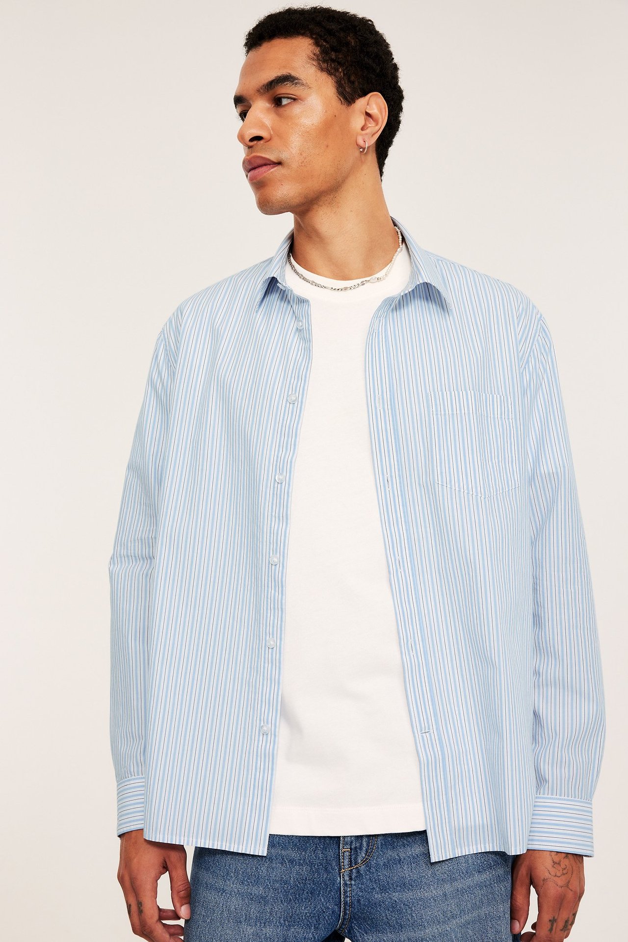 Widok z przodu/boku: Mężczyzna w rozpiętej błękitnej koszuli w paski, białym t-shircie i niebieskich dżinsach.