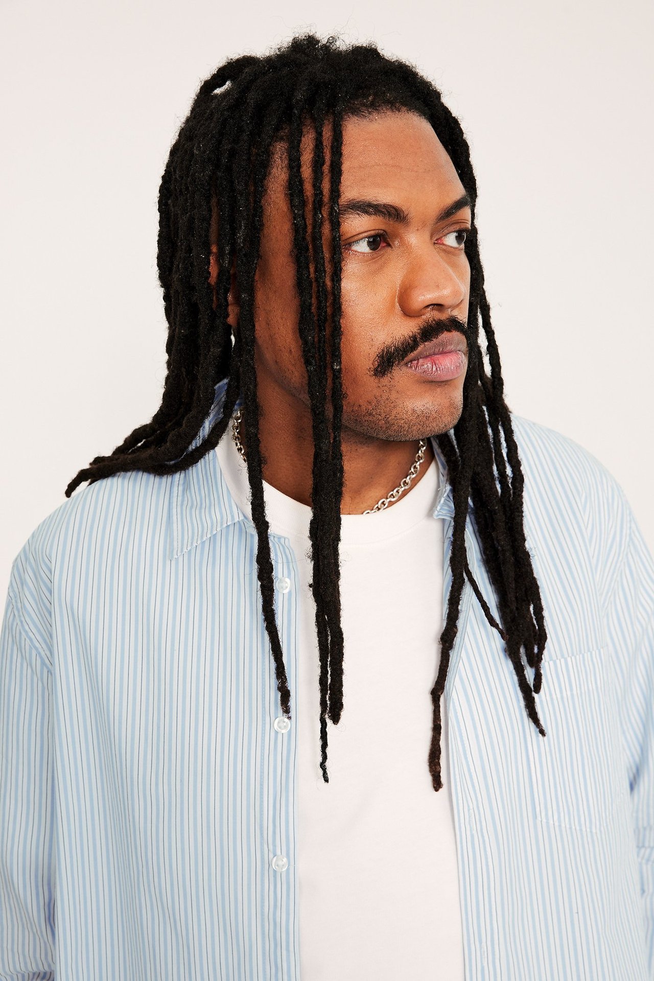 Lähikuva miehestä vaaleansinisessä raidallisessa popliinipaidassa valkoisen t-paidan ja hopeakaulakorun päällä.