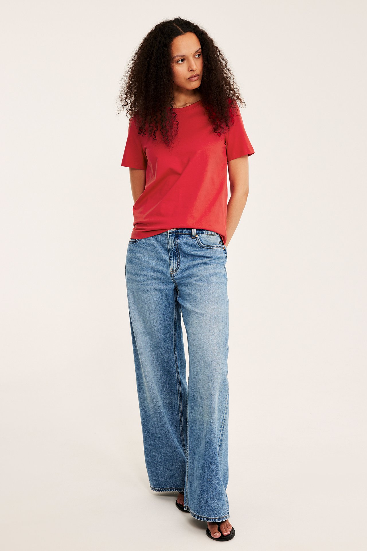 Edestä naisesta punaisessa puuvilla T-paidassa, sinisissä leveälahkeisissa farkuissa ja mustissa varvastossuissa.