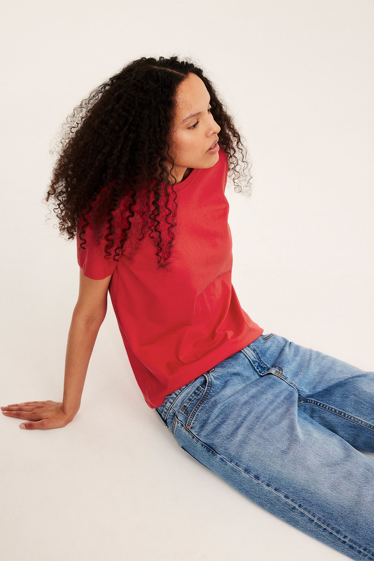 Sideprofil av en kvinne i en rød, rett passform, kortermet bomulls-T-skjorte og lyseblå jeans.