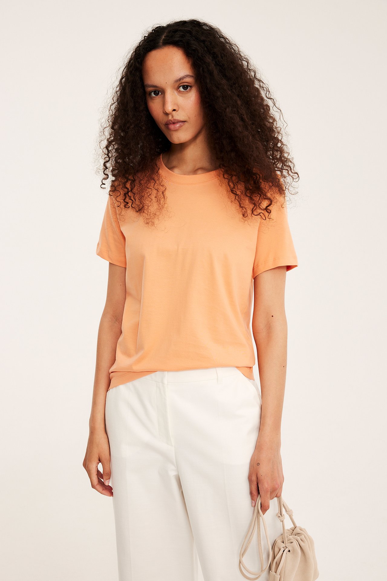 Framifrån: kvinna i orange rak bomulls-t-shirt, vita byxor, håller beige handväska.