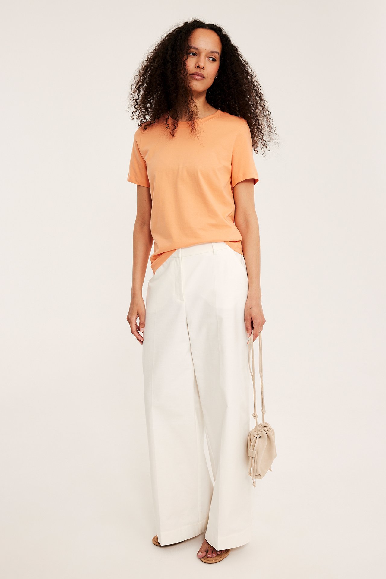 Frontvy: kvinna i orange T-shirt, vita vida byxor, sandaler, håller beige väska.