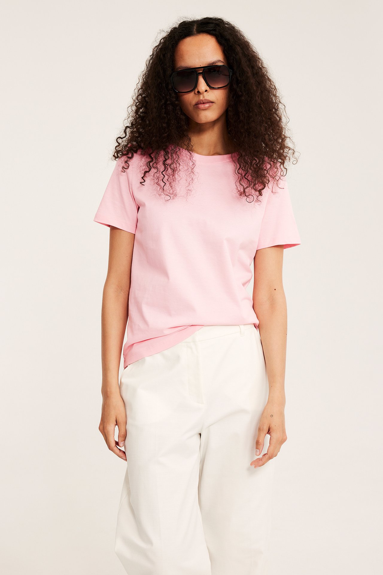 Naisen etukuva vaaleanpunaisessa puuvilla-T-paidassa, valkoisissa housuissa ja mustissa pilotsolmulaseissa.