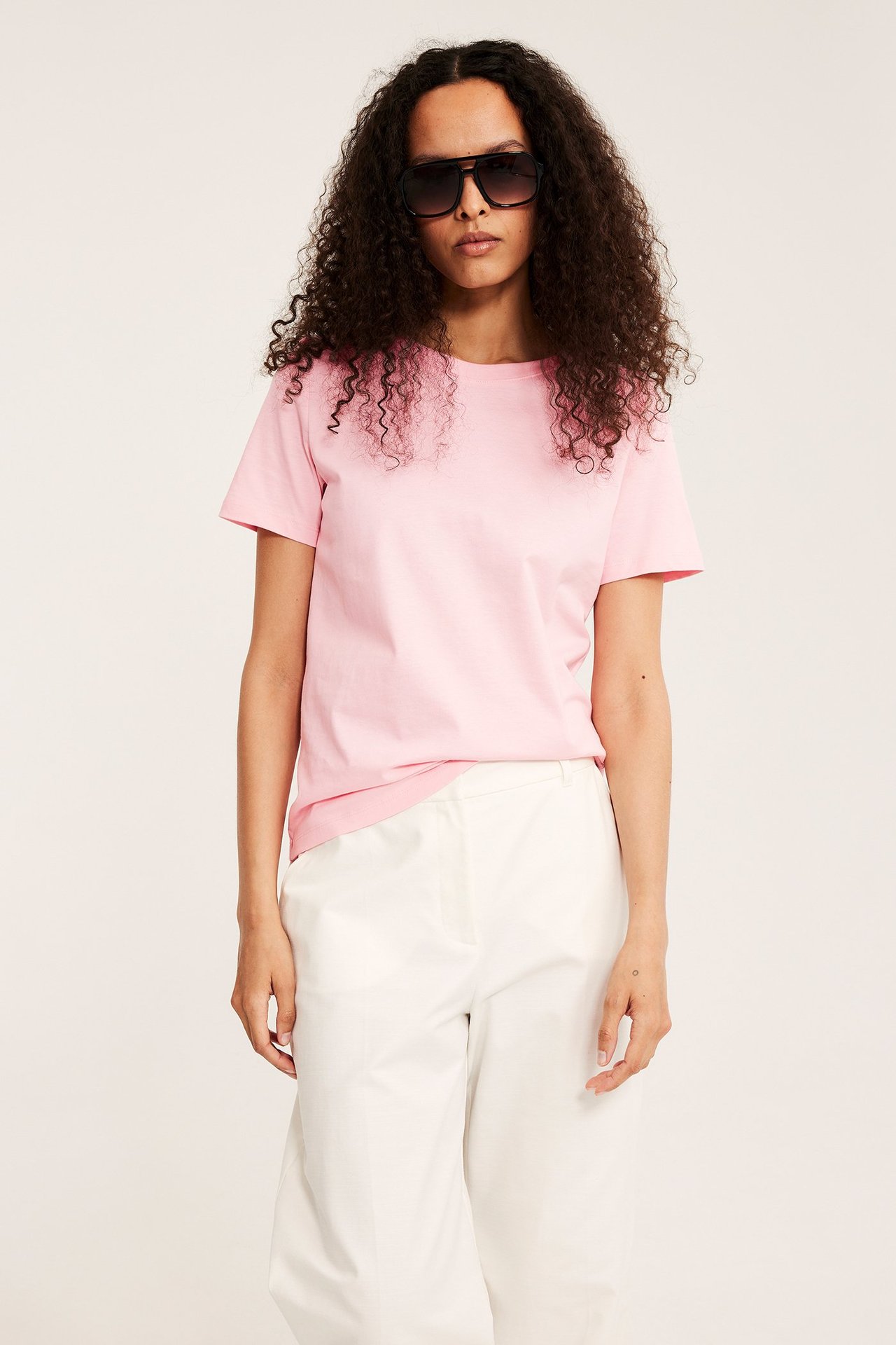 Widok z przodu kobiety w jasnoróżowej bawełnianej koszulce, białych spodniach i czarnych okularach aviator.