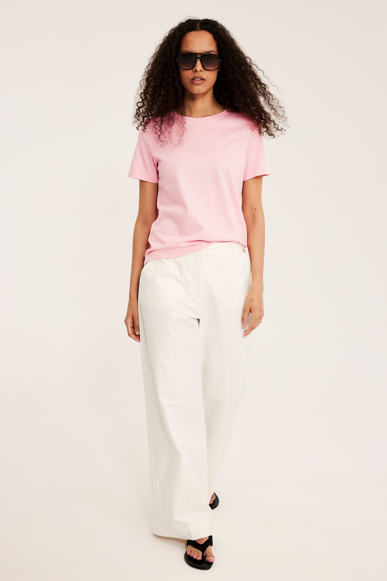 Edestäpäin, nainen yllään vaaleanpunainen puuvilla-T-paita, valkoiset leveälahkeiset housut, mustat aurinkolasit ja sandaalit.