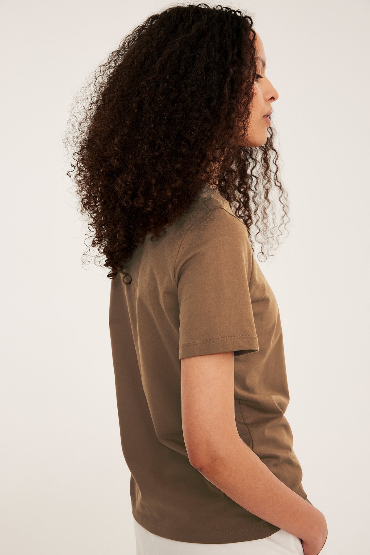 Sivukuva naisesta ruskeassa, lyhythihaisessa, suoraleikkauksisessa puuvilla T-paidassa ja valkoisessa alaosassa.