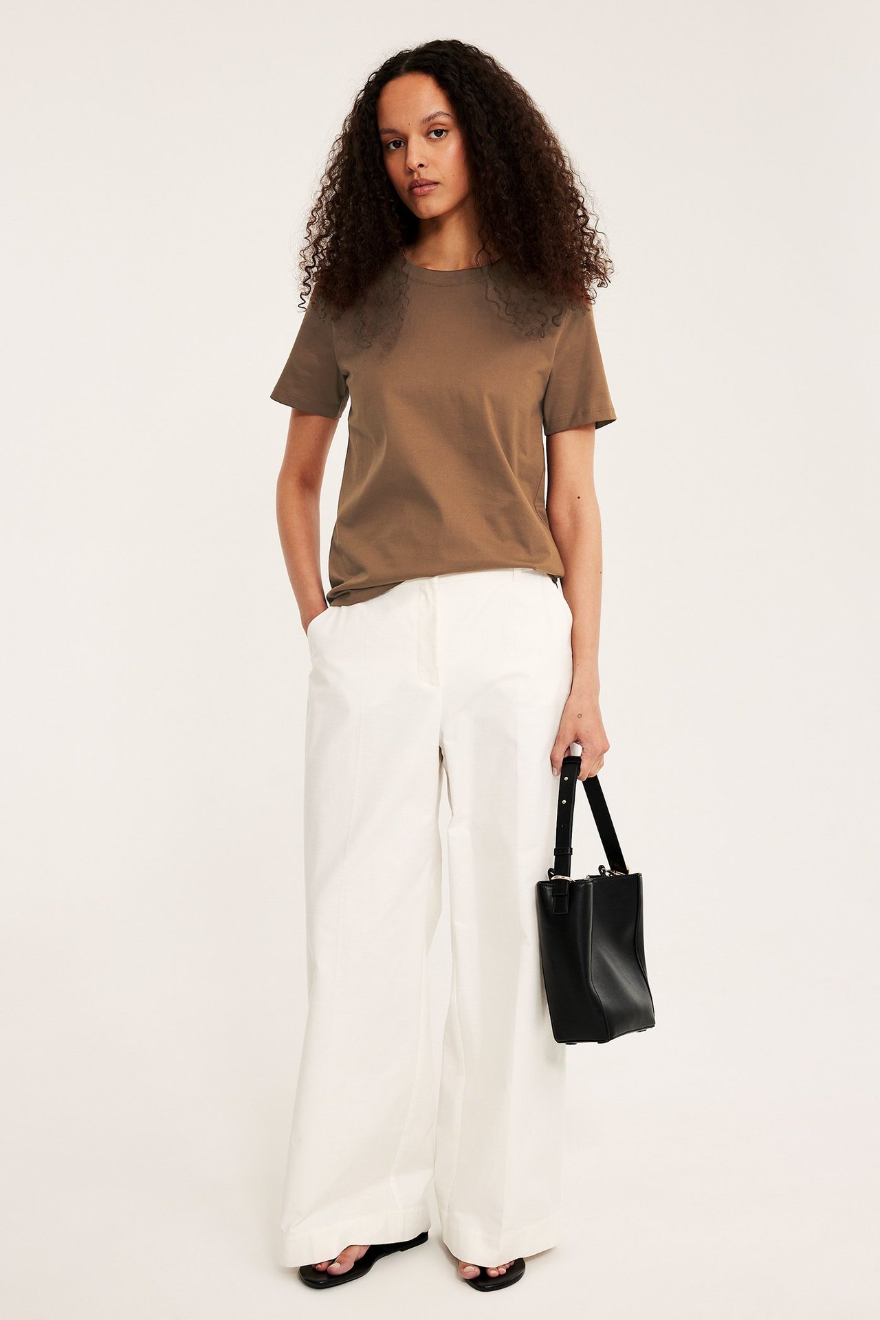 Nainen edestä: Ruskea puuvilla-T-paita, valkoiset leveät housut, mustat sandaalit, musta käsilaukku.