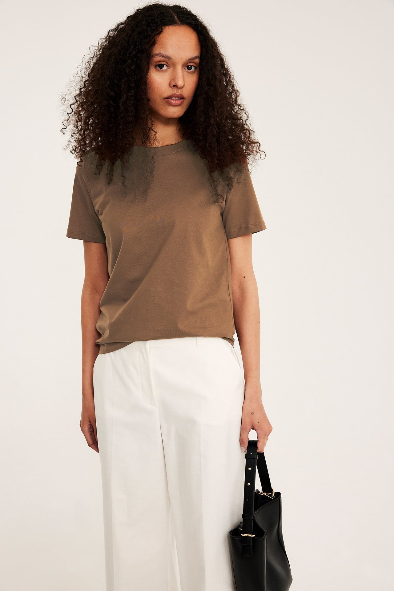 Naisen etukuva ruskeassa suorassa puuvilla-T-paidassa, valkoisissa leveissä housuissa, mustaa laukkua kantaen.