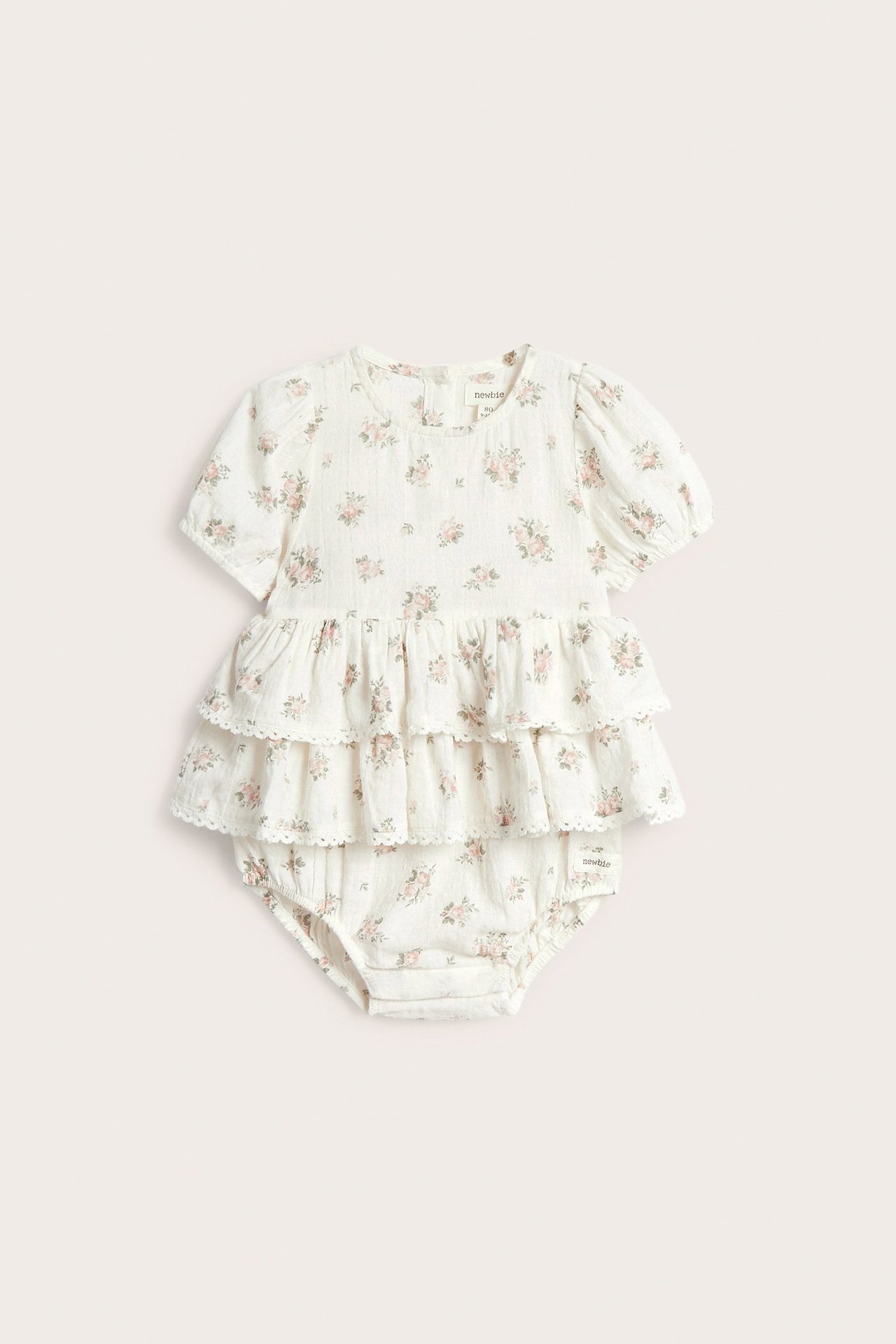 Forfra, hvit babybomullsdress med blomstermønster, puffermer og volanger.