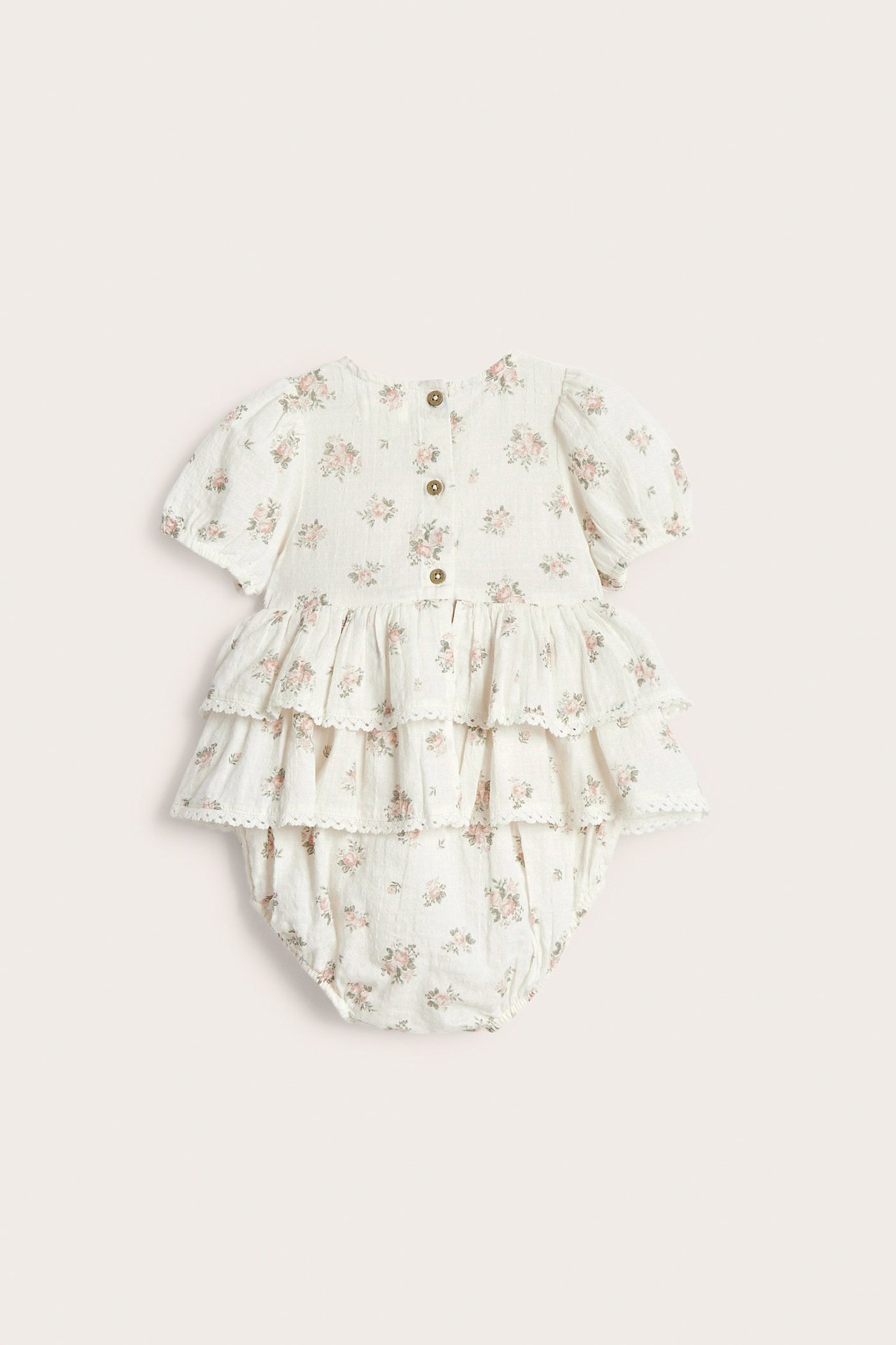 Baksidebilde av en offwhite baby jumpsuit i bomull med rosa blomstertrykk, volanger, blonder og knapper.