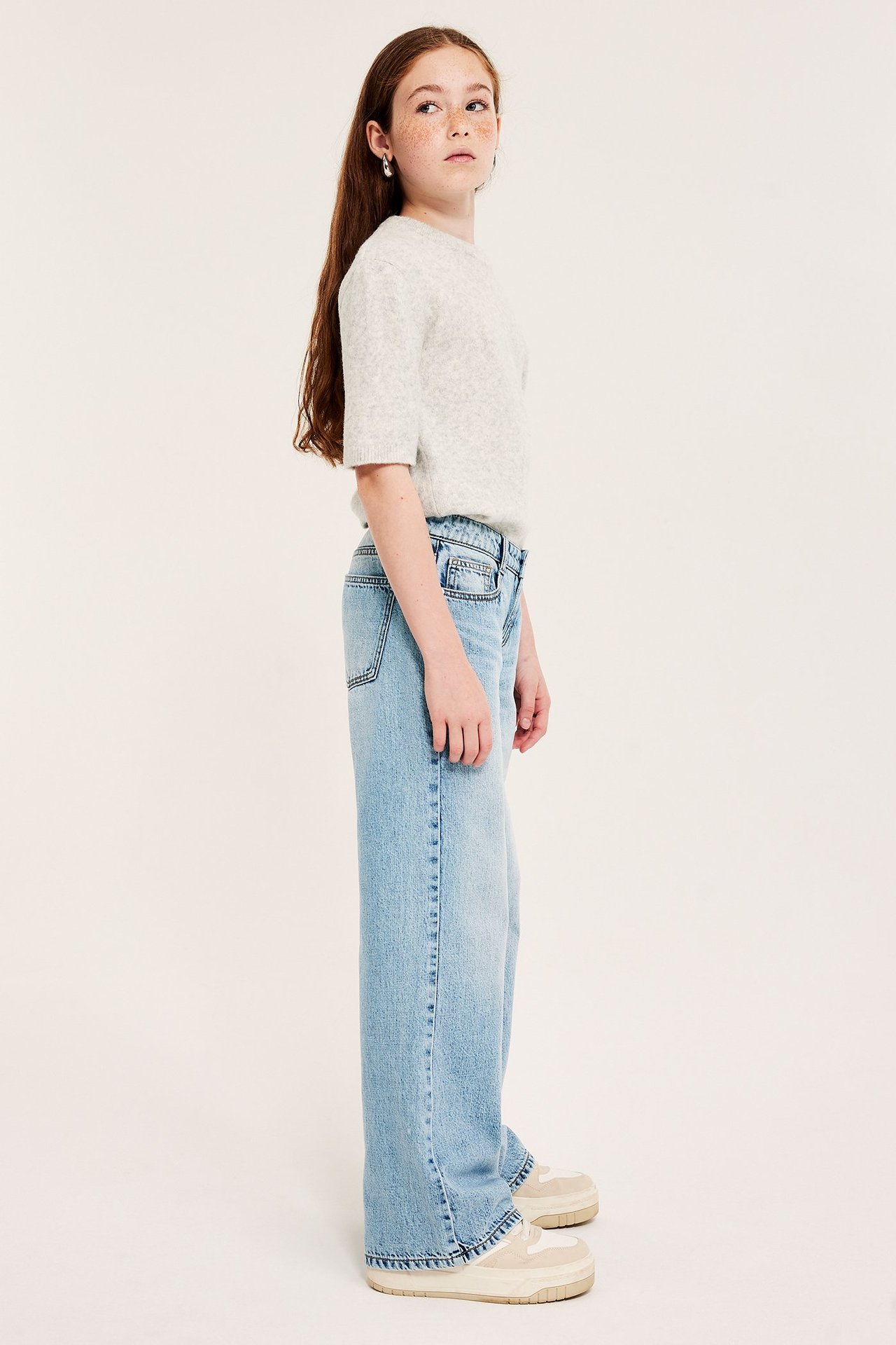 Sivukuva lapsesta vaaleissa leveissä matalavyötäröfarkuissa, vaaleanharmaassa topissa ja tennareissa.