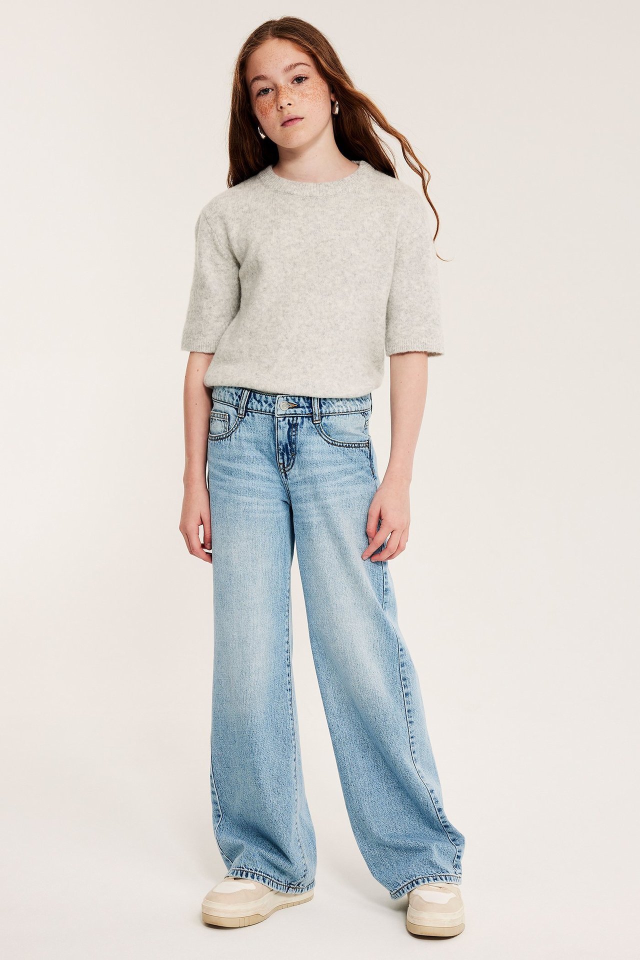 Lapsi edestä: vaaleat leveät farkut, vaaleanharmaa lyhythihainen neule ja beiget tennarit.