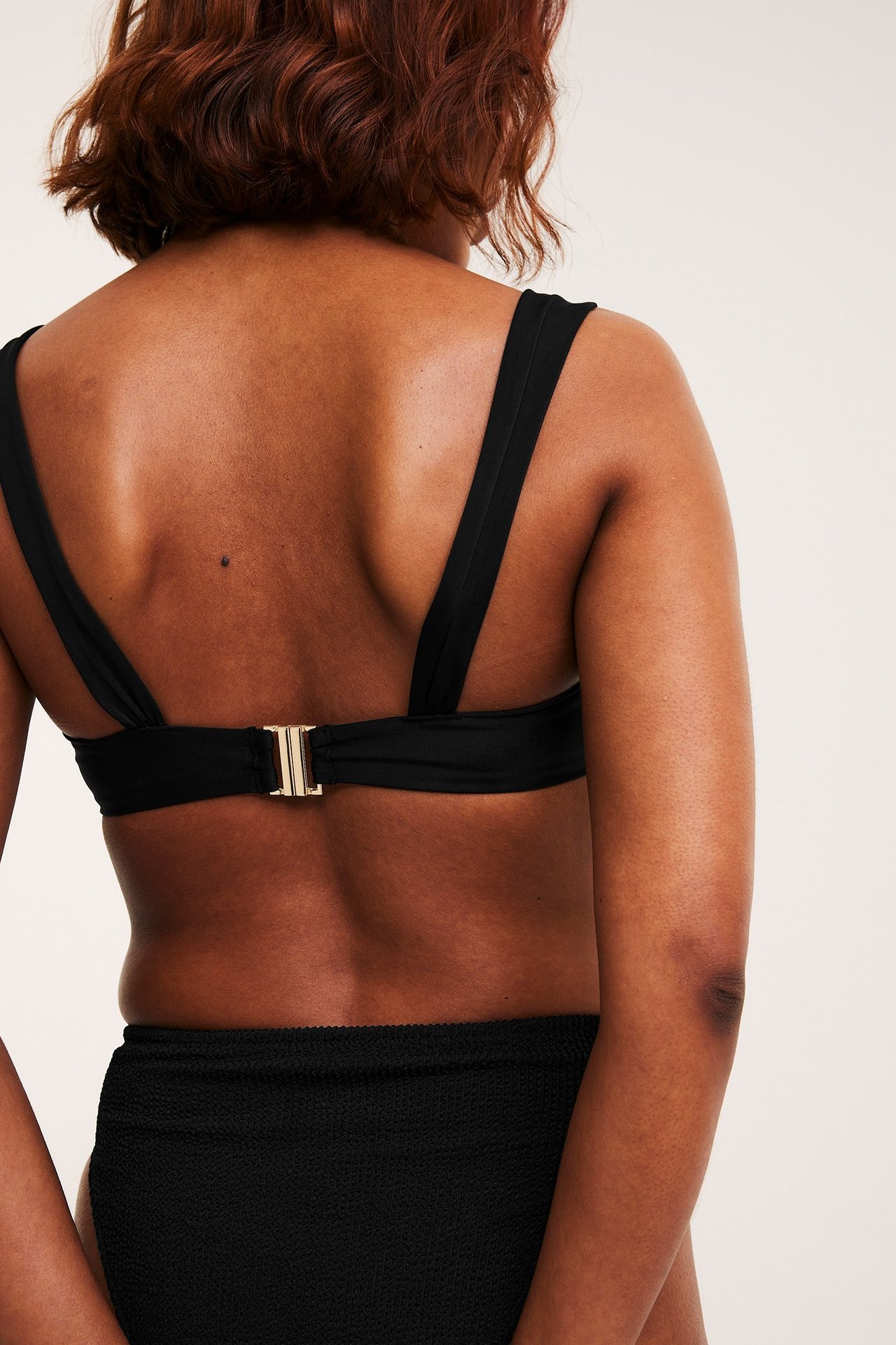 Takaa: nainen mustassa kaarituellisessa bikinirintaliivissä leveillä olkaimilla, kultaisella soljella sekä mustilla alaosilla.