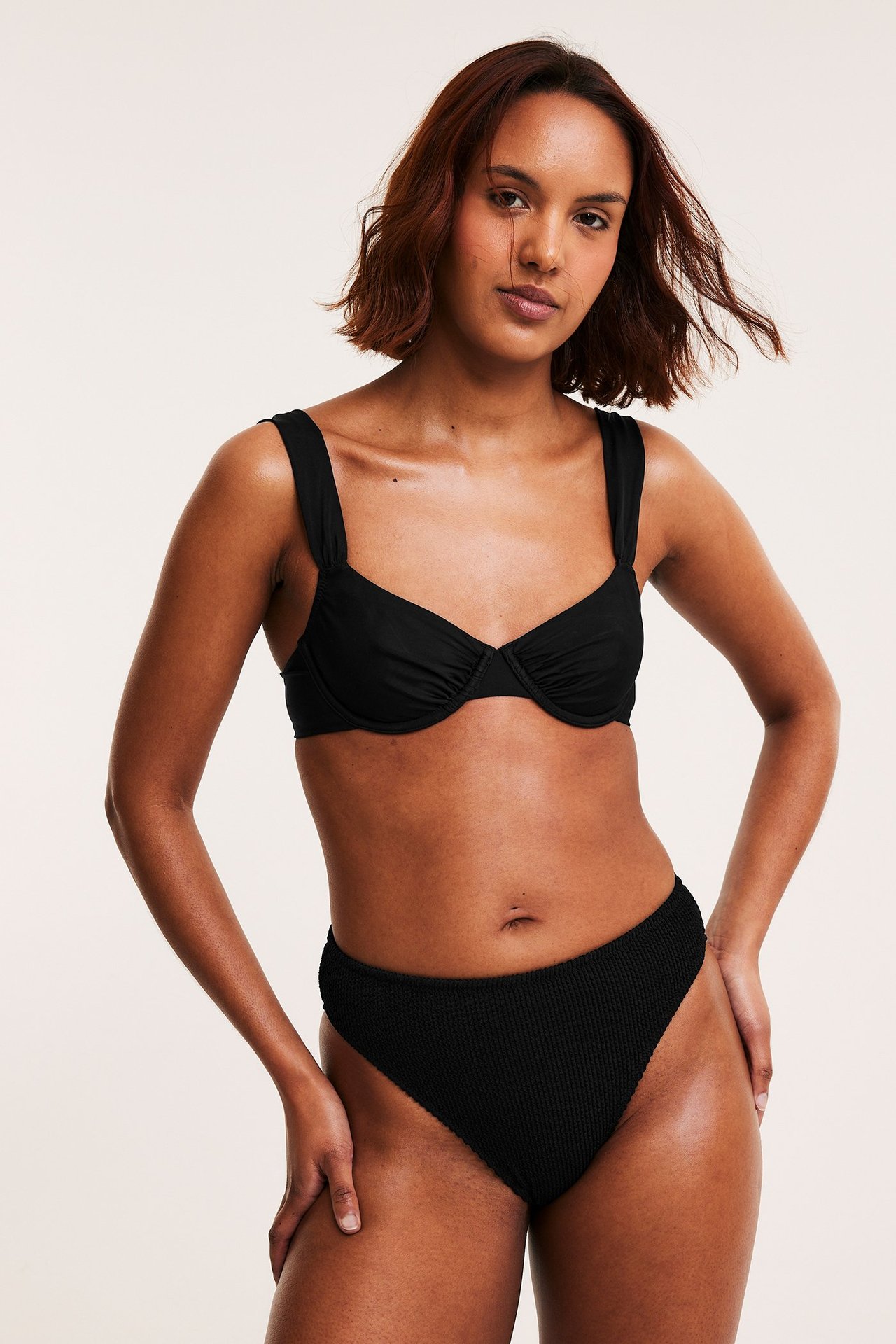 Musta kaarituellinen bikinirintaliivi leveillä olkaimilla ja mustat kuvioidut bikinialaosat, naisen yllä.