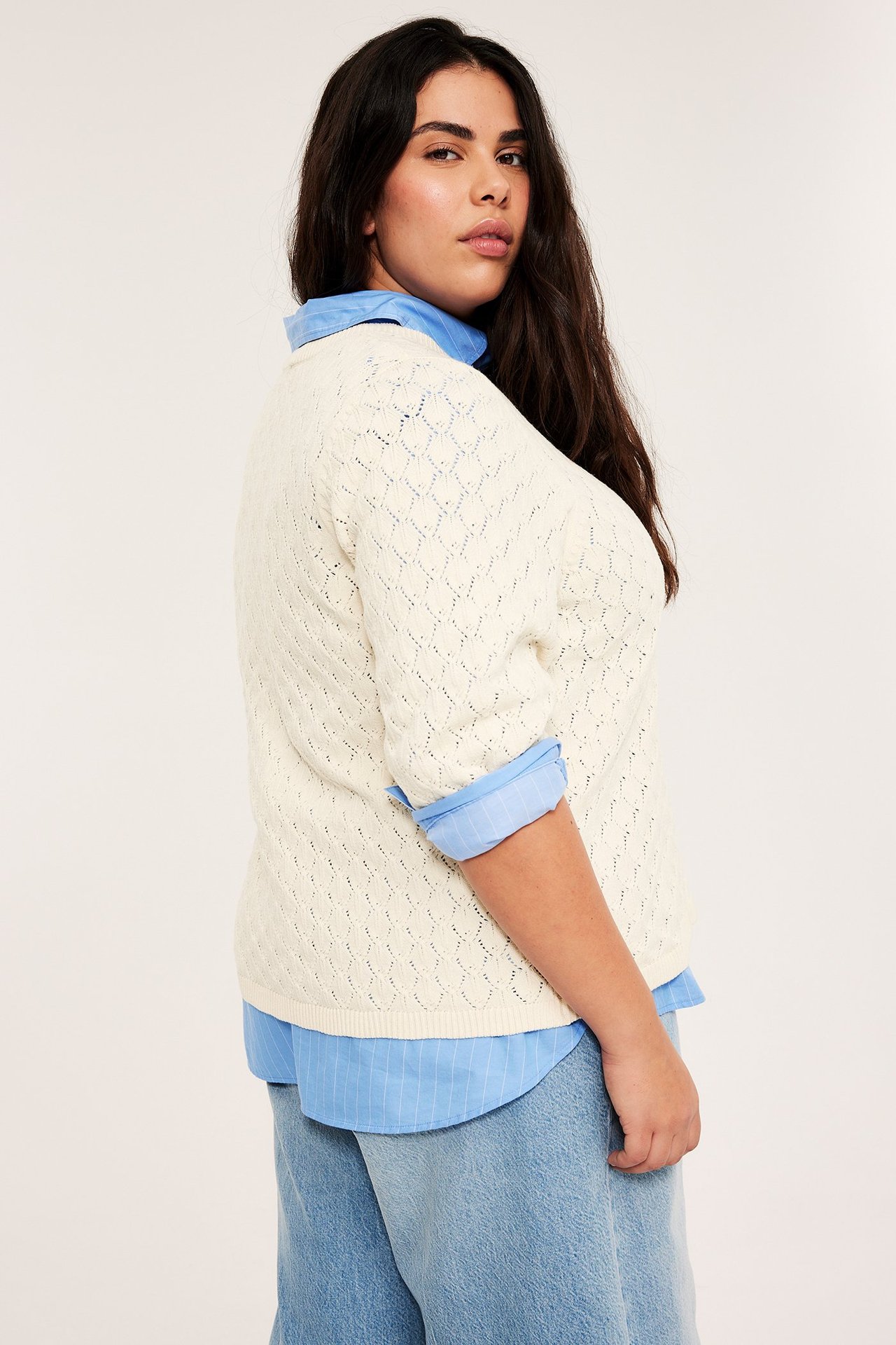 Sidovy av kvinna i offwhite spetsstickad kortärmad tröja över blårandig skjorta och ljusblå jeans.