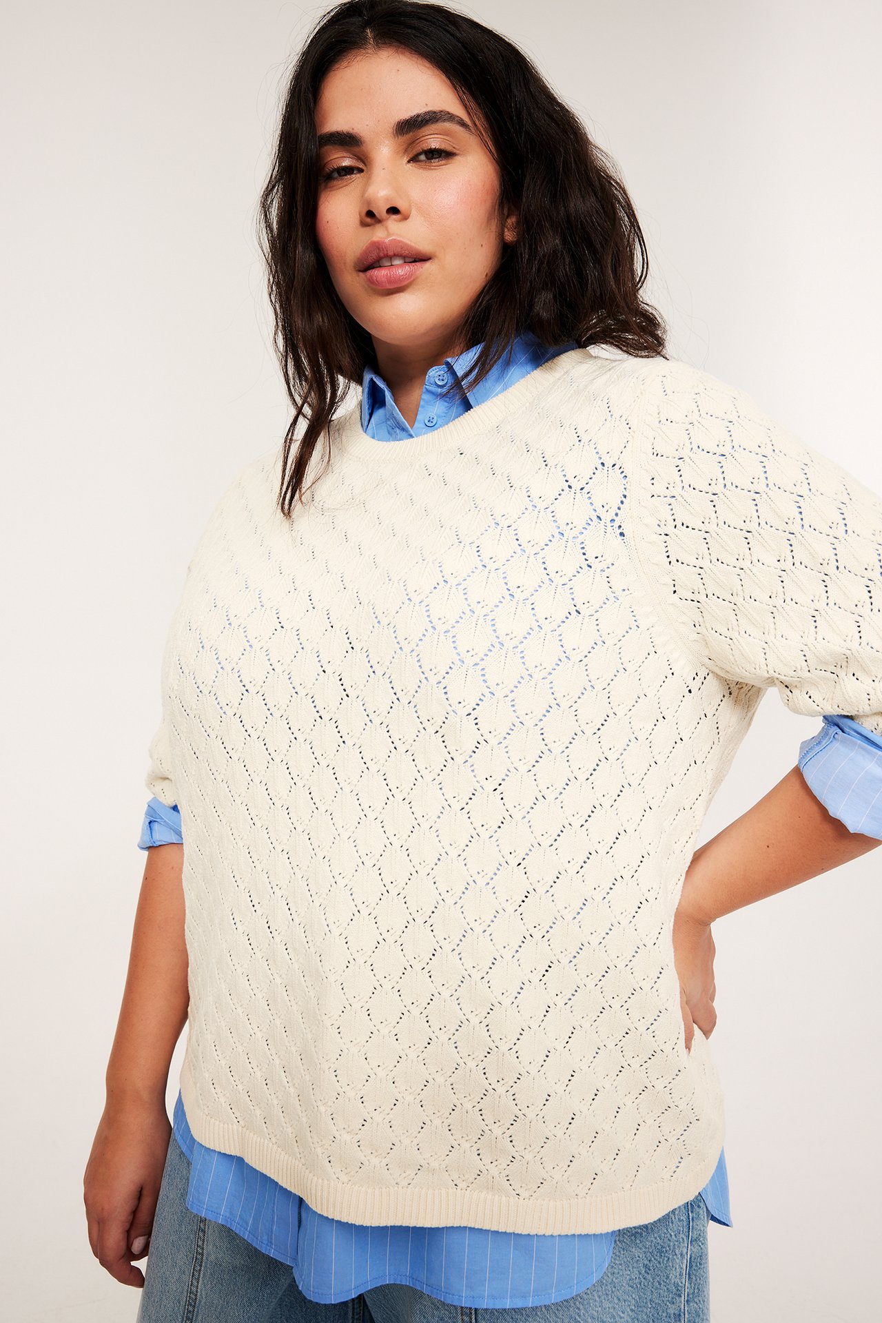 Edestä kuvattu nainen yllään luonnonvalkoinen pitsineulettu lyhythihainen pusero sinisen paidan ja farkkujen päällä.