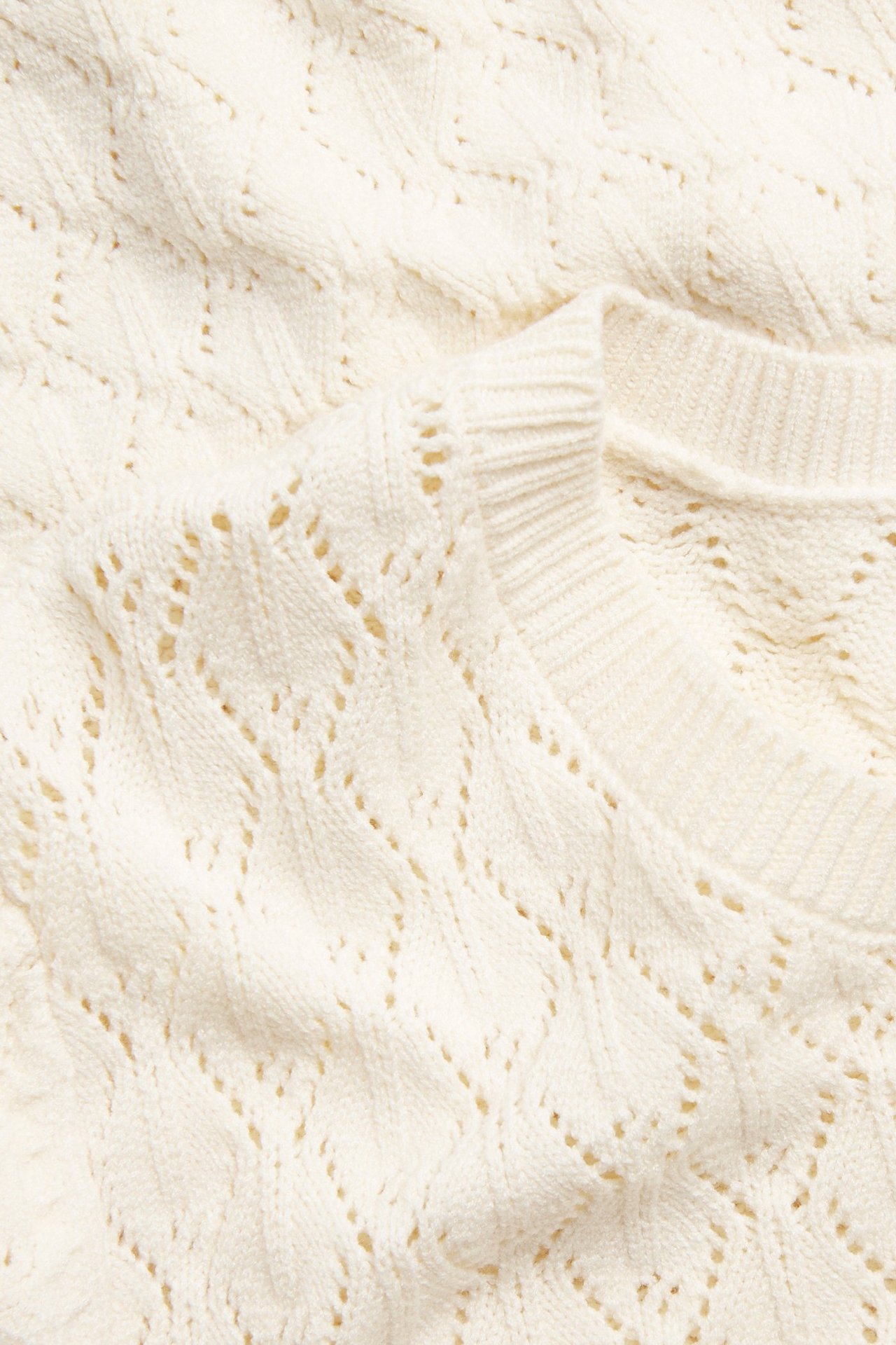 Zbliżenie na ażurowy sweter w kolorze off-white z prążkowanym okrągłym dekoltem.