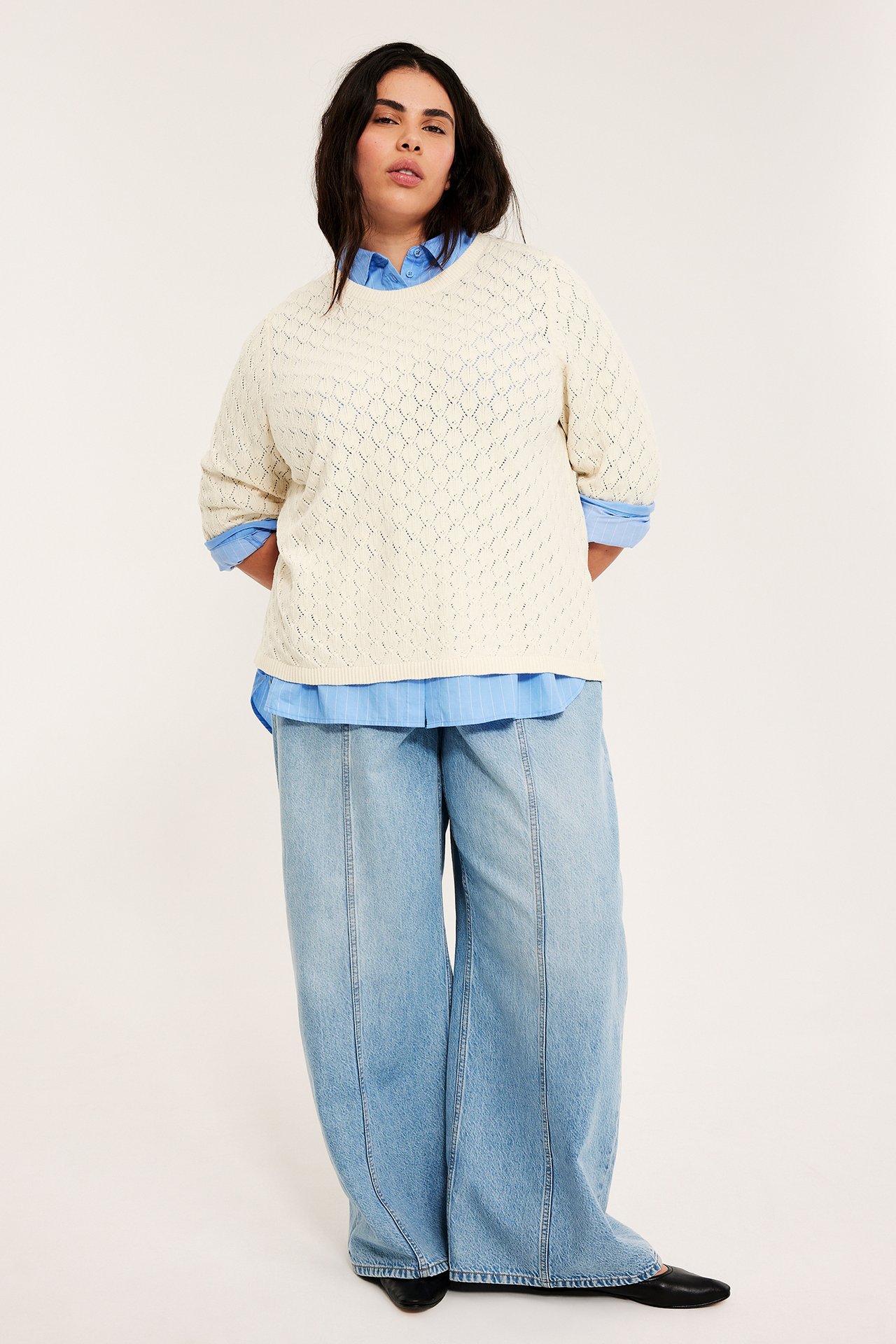 Widok z przodu: Kobieta w kremowym koronkowym swetrze z krótkim rękawem, na jasnoniebieskiej koszuli i luźnych dżinsach.