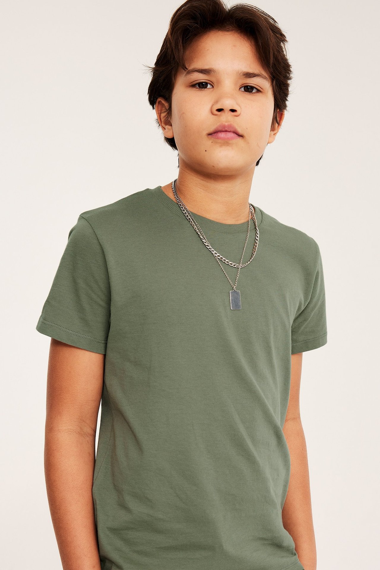 Edestä, lapsi yllään yksivärinen vihreä puuvillainen T-paita ja hopeinen kerroksellinen kaulakoru.