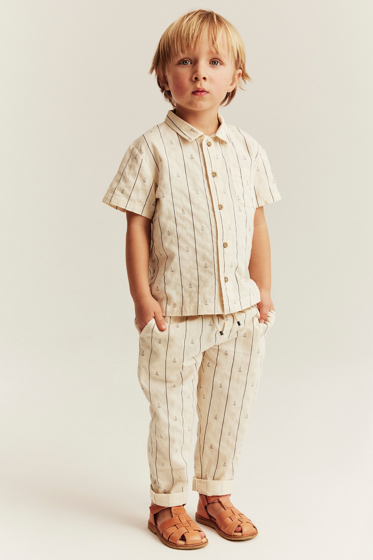 Frontvy: Pojke i benvit vävd kortärmad skjorta med segelbåtsränder, matchande byxor, bruna sandaler.